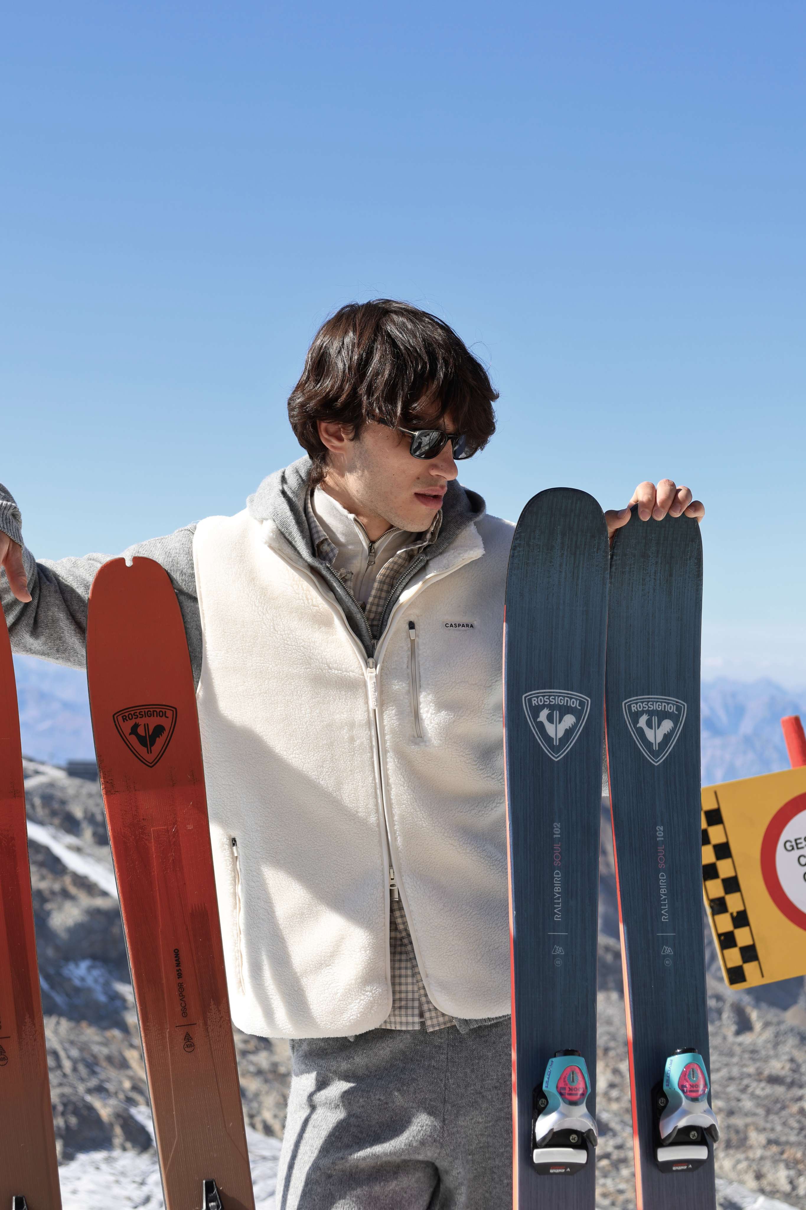 Man wearing a creme CASPARA Cloudfleece vest holding skis on a snowy mountain.