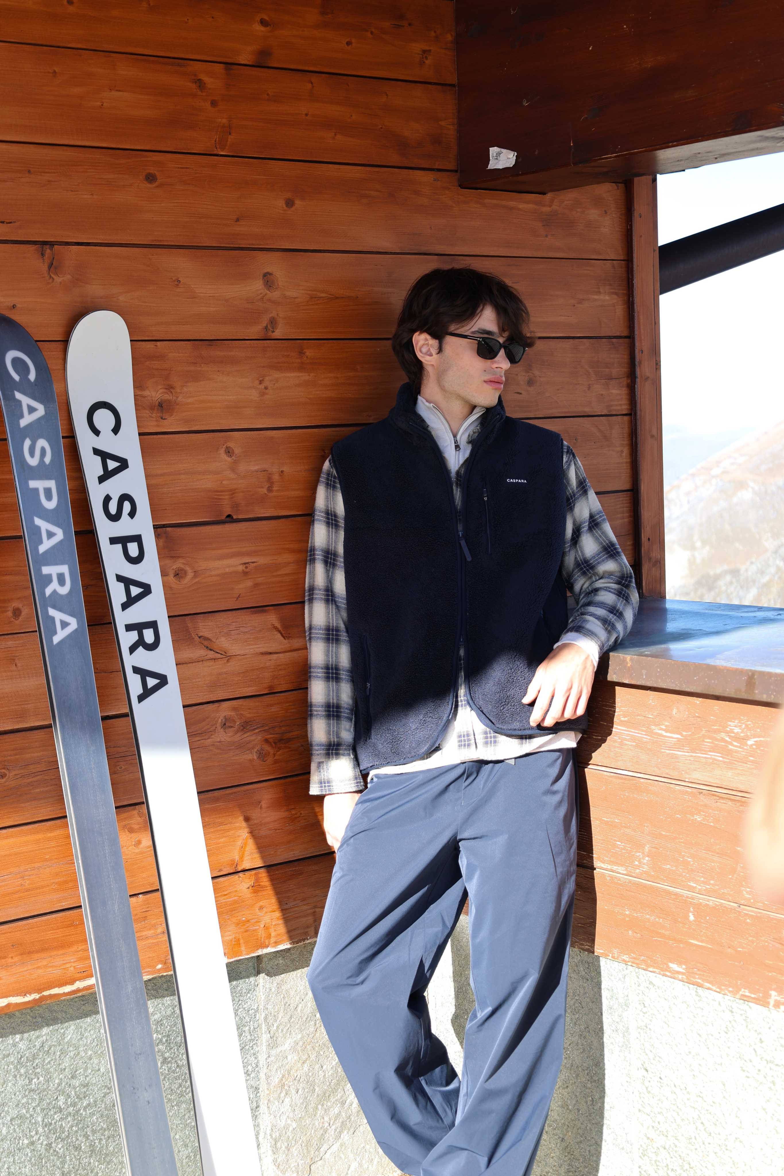 Man wearing a navy CASPARA Cloudfleece vest standing next to skis by a wooden chalet.