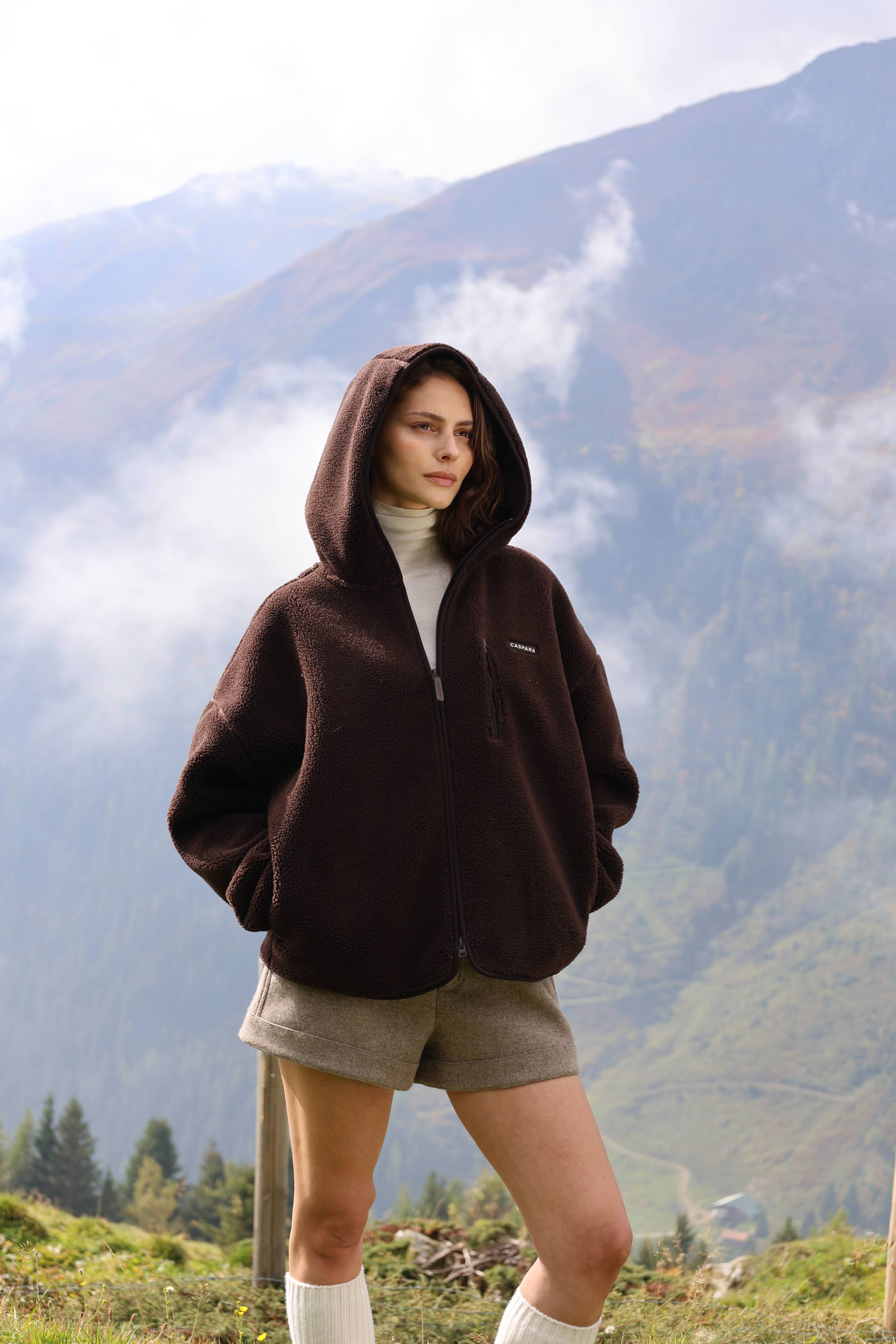 Woman wearing an espresso CASPARA Cloudfleece hooded jacket outdoors in the mountains.