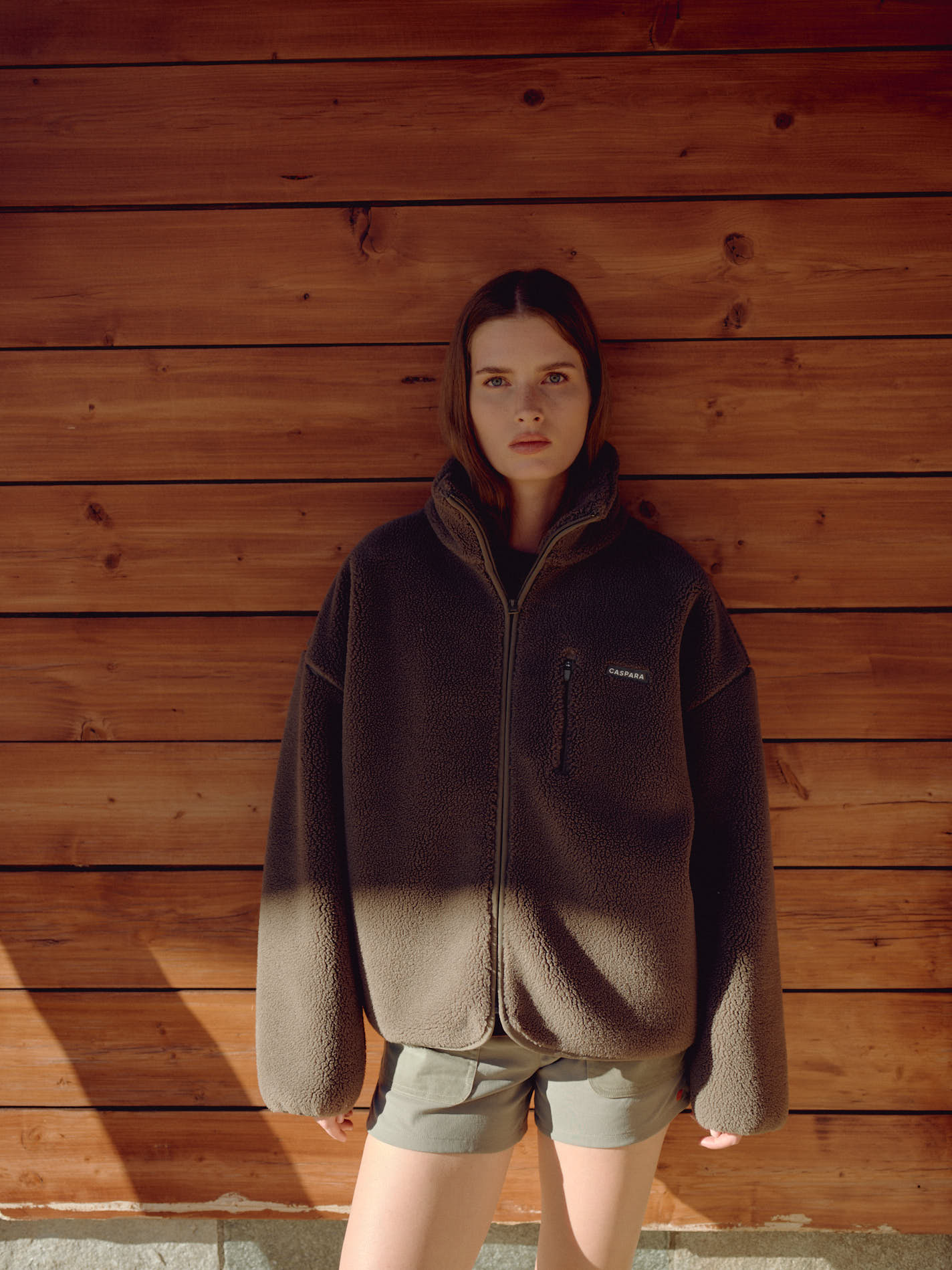 Woman wearing a khaki CASPARA Cloudfleece jacket standing against a wooden wall.