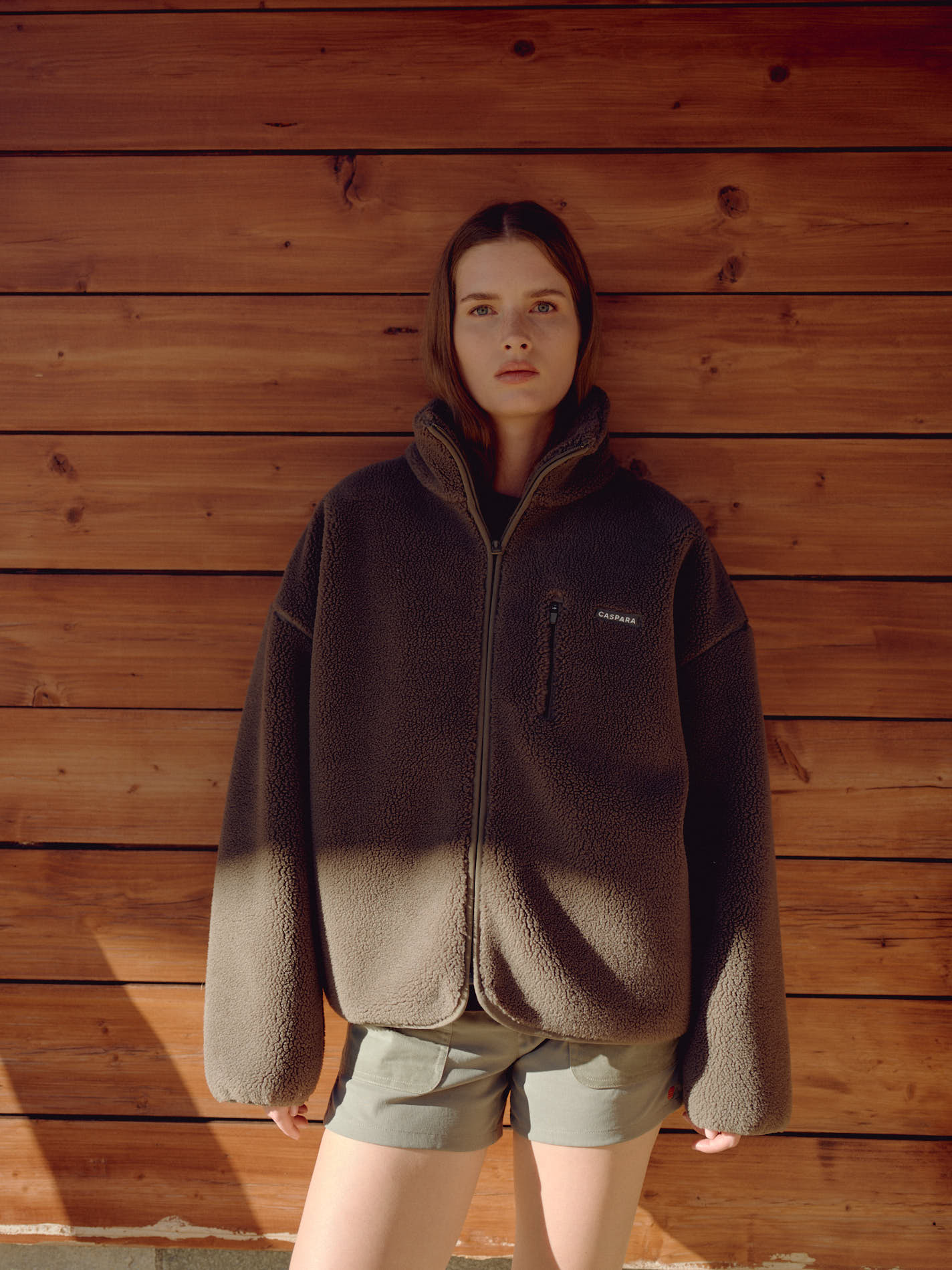 Woman wearing an oversized khaki CASPARA Cloudfleece jacket against a wooden wall.