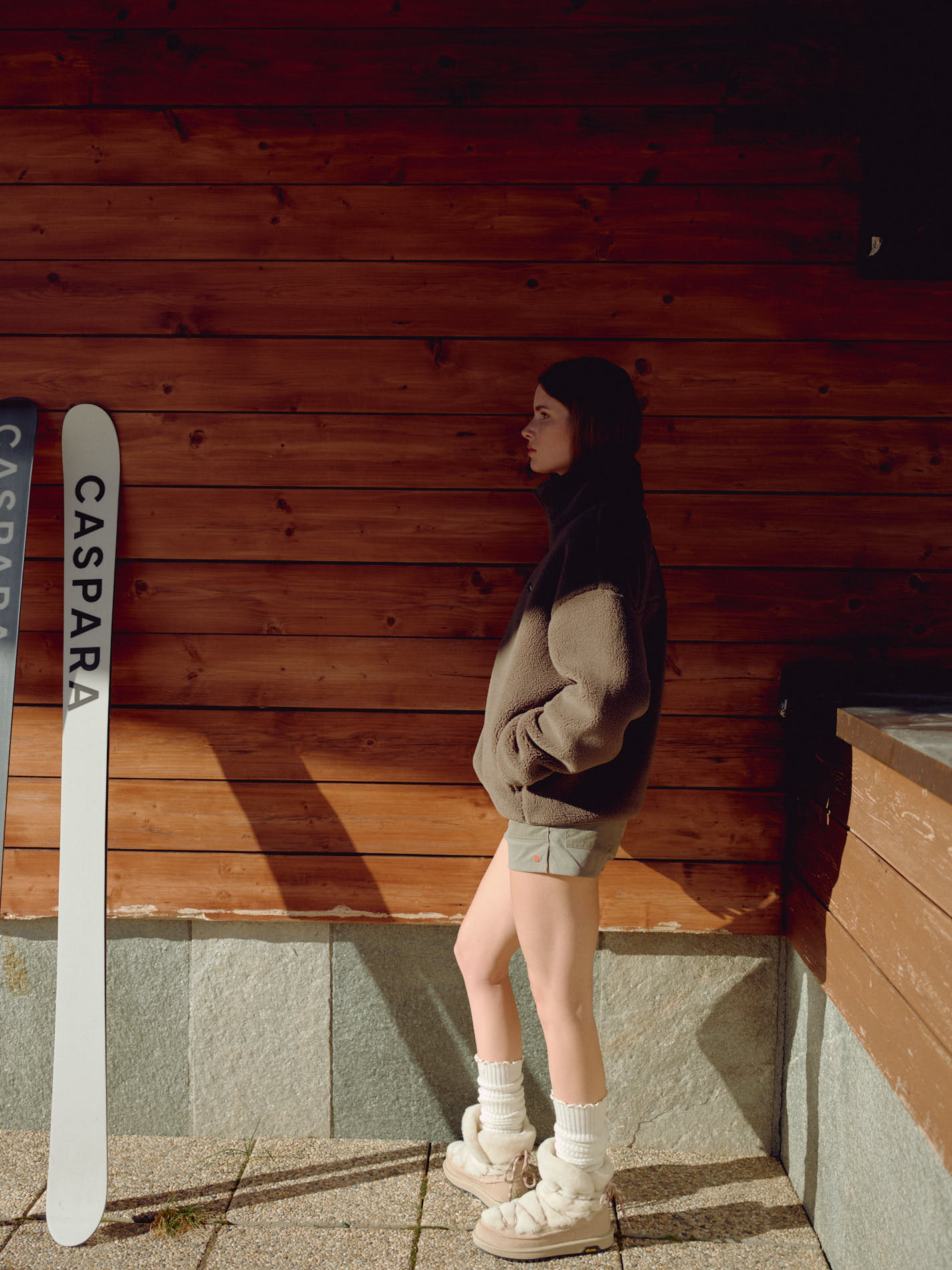 Woman wearing a khaki CASPARA Cloudfleece jacket standing next to skis by a wooden wall.