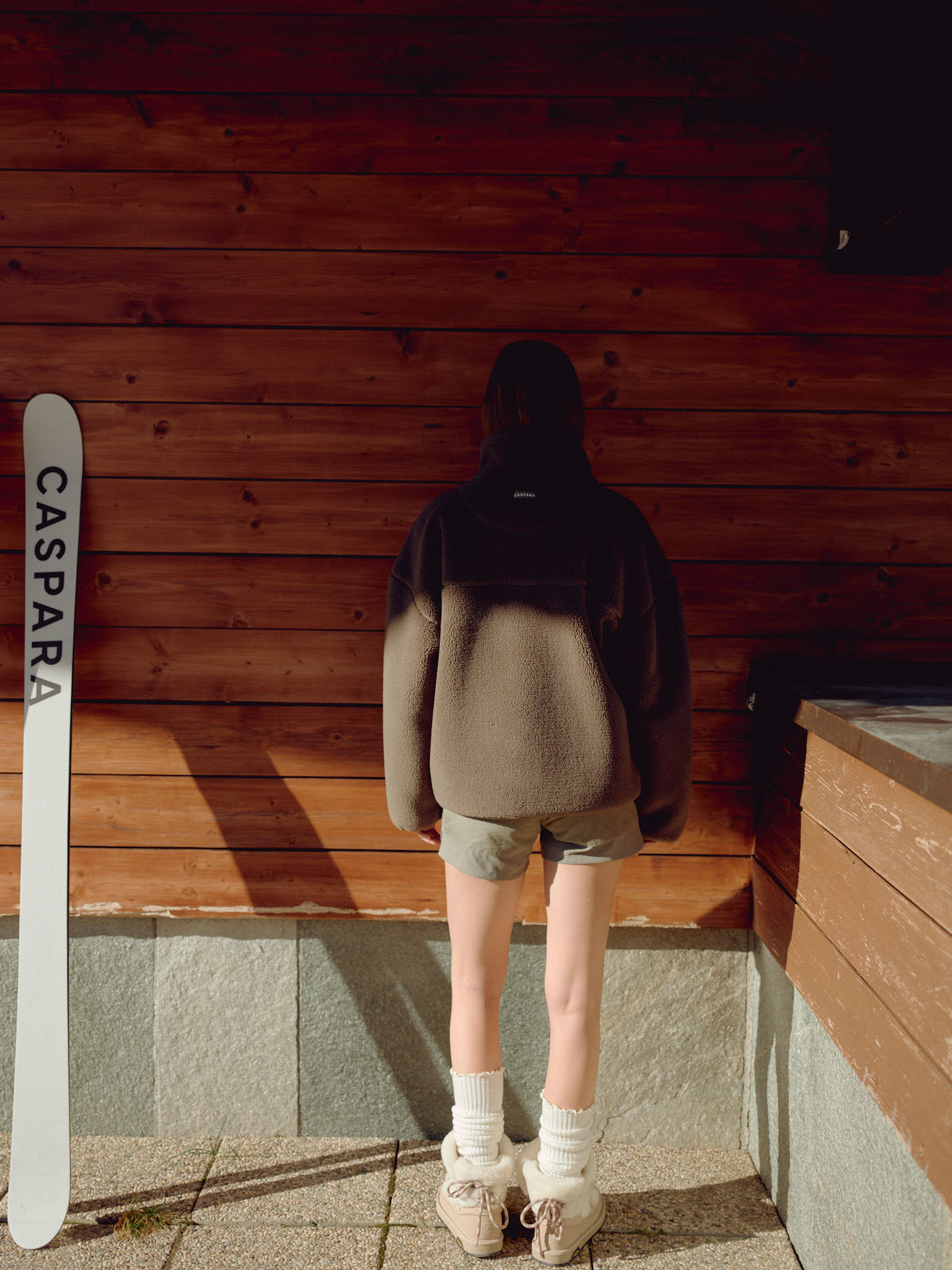 Back view of a woman wearing a khaki CASPARA Cloudfleece jacket against a wooden wall.
