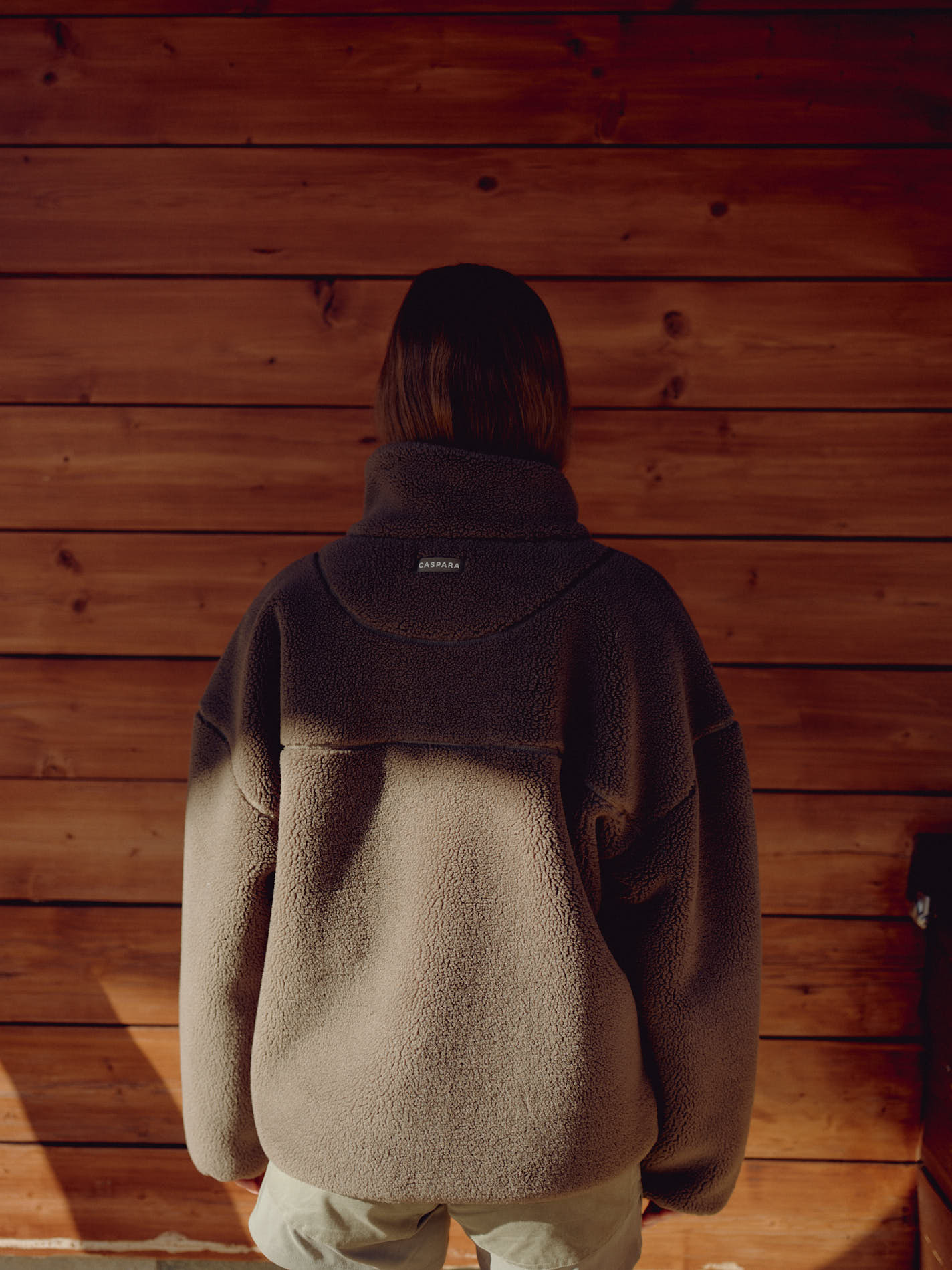 Back view of woman wearing a khaki CASPARA Cloudfleece jacket against a wooden wall.
