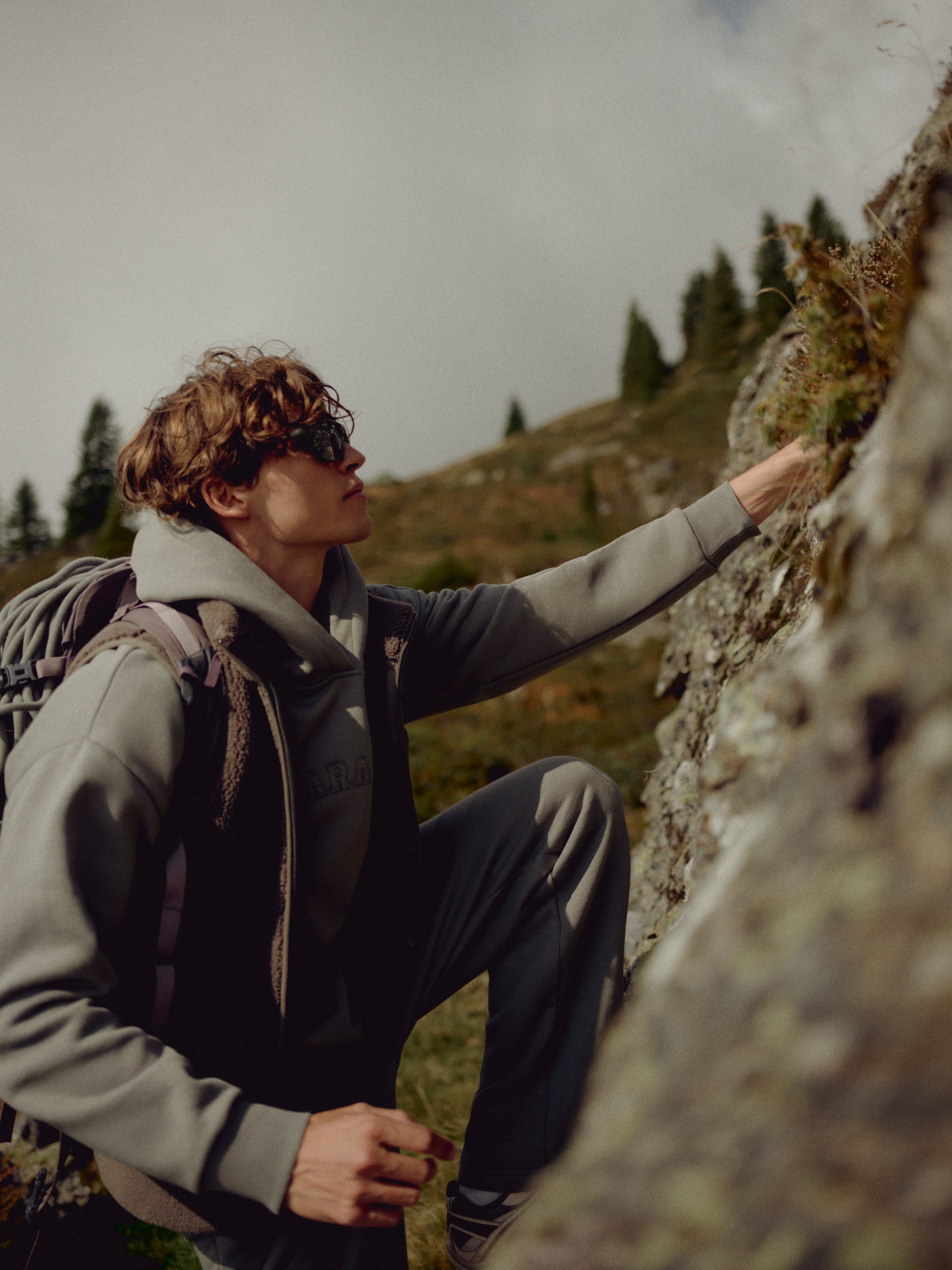 Man wearing a khaki CASPARA CS hoodie outdoors on a rocky mountain slope.