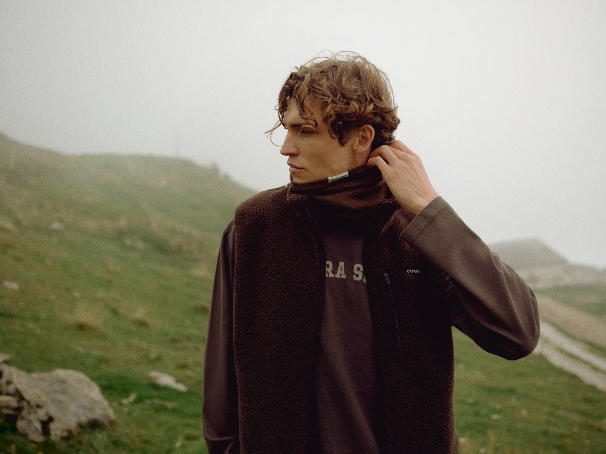 Man wearing an espresso CASPARA Cloudfleece vest in a misty mountain landscape.