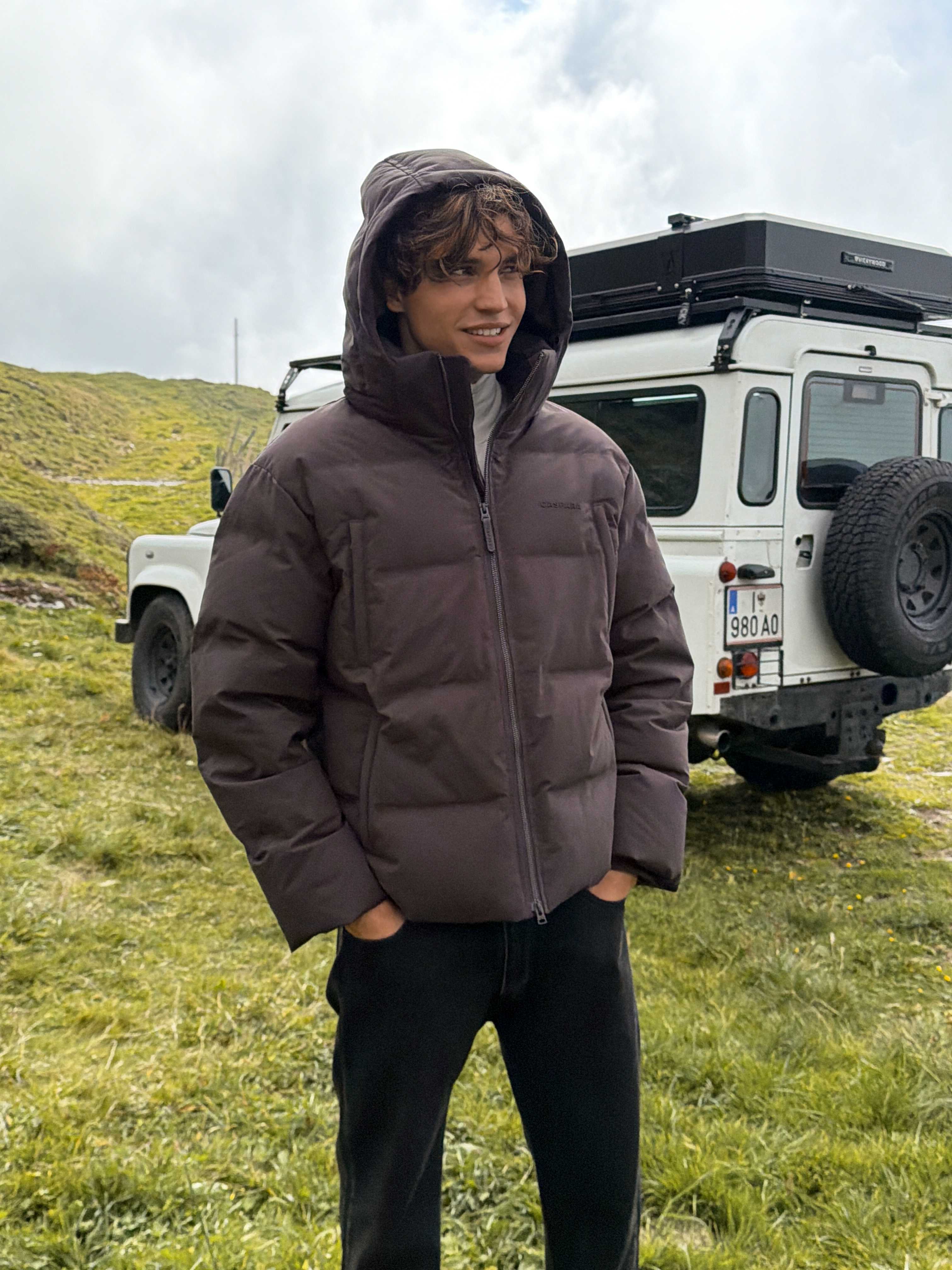 Man wearing a cocoa bean CASPARA Boreal puffer jacket outdoors in front of an off-road vehicle.