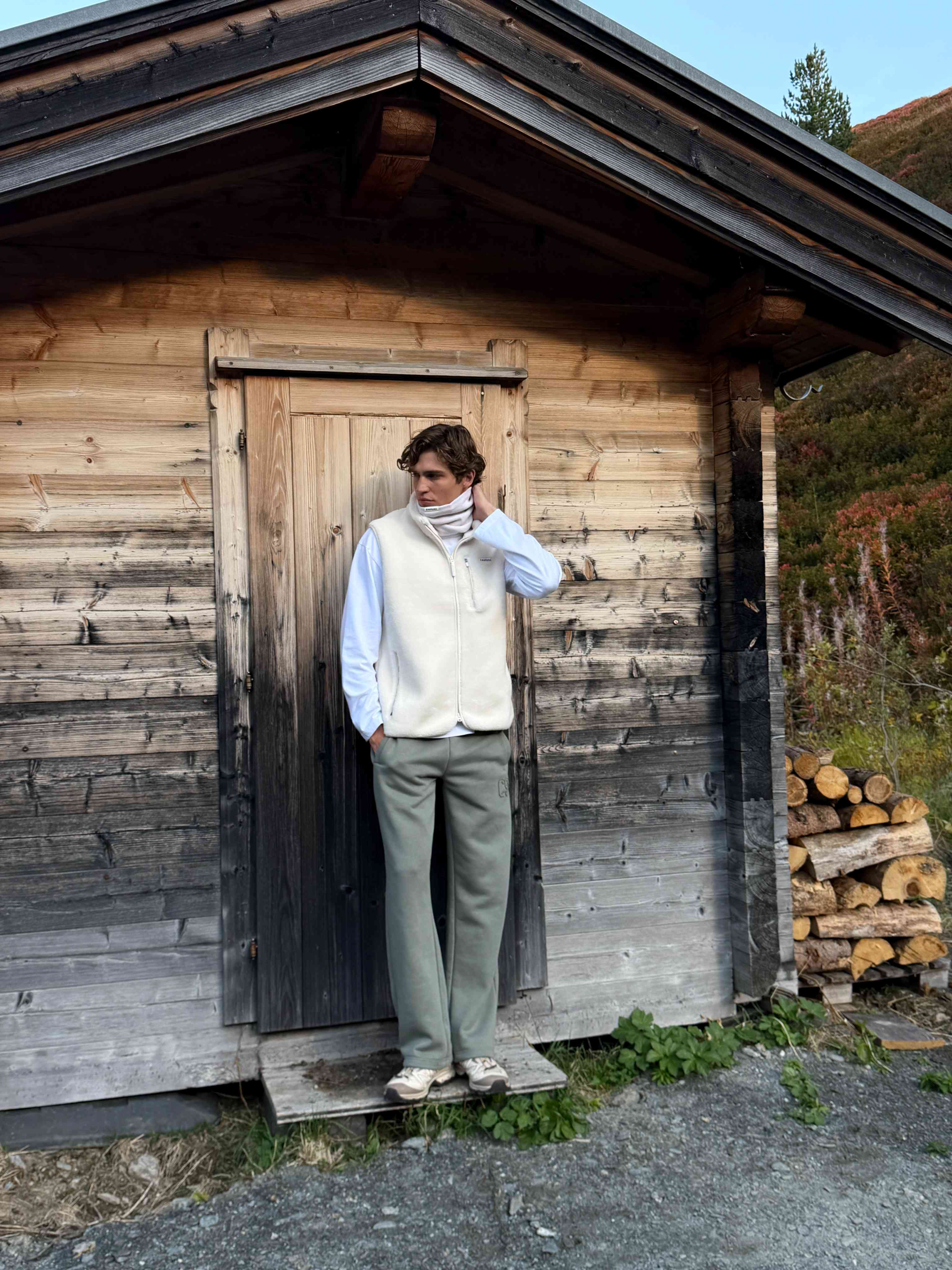 Man wearing a creme CASPARA Cloudfleece vest outside a rustic wooden cabin.