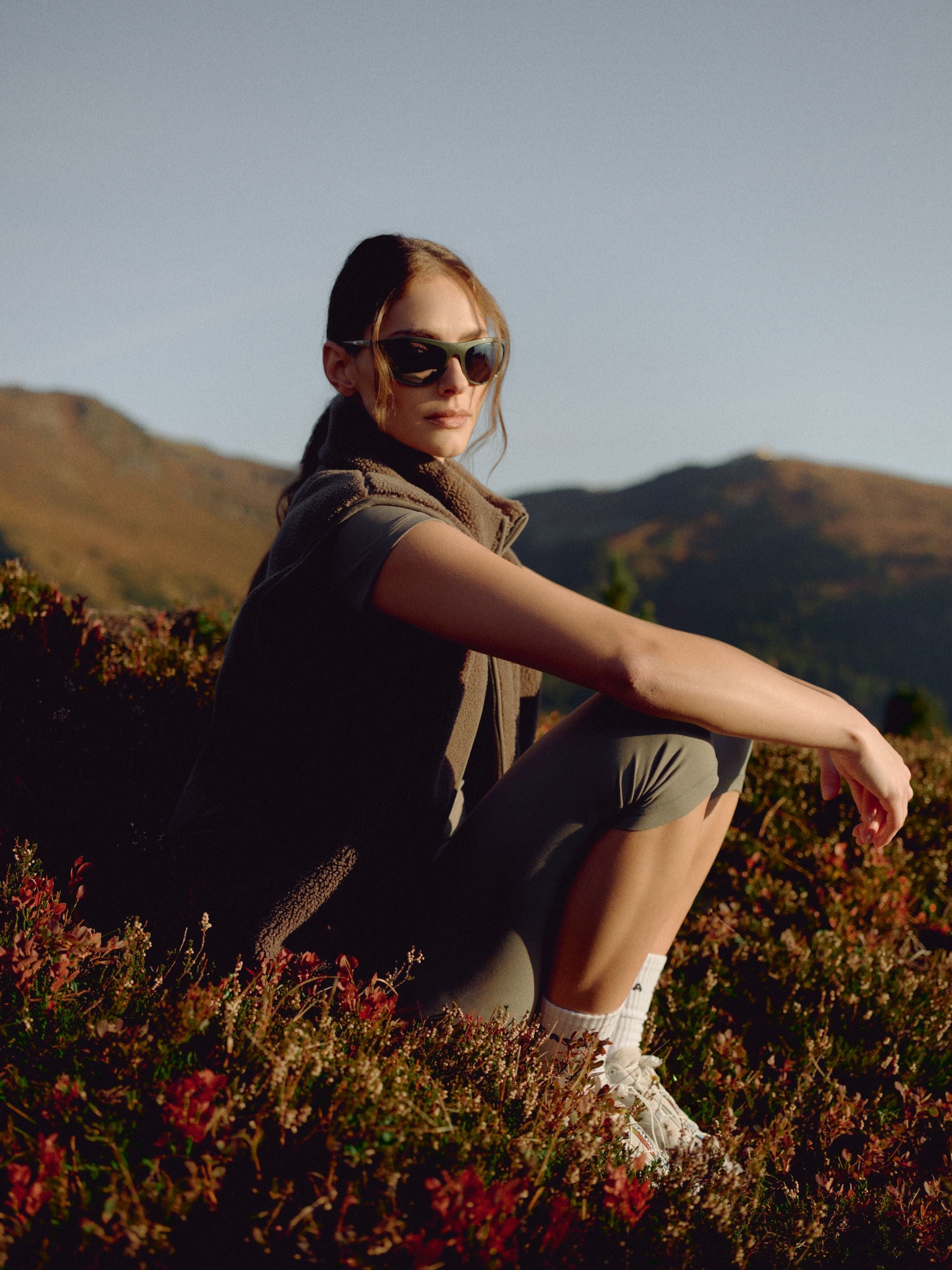 Woman wearing a khaki CASPARA Cloudfleece vest sitting in a mountain landscape.