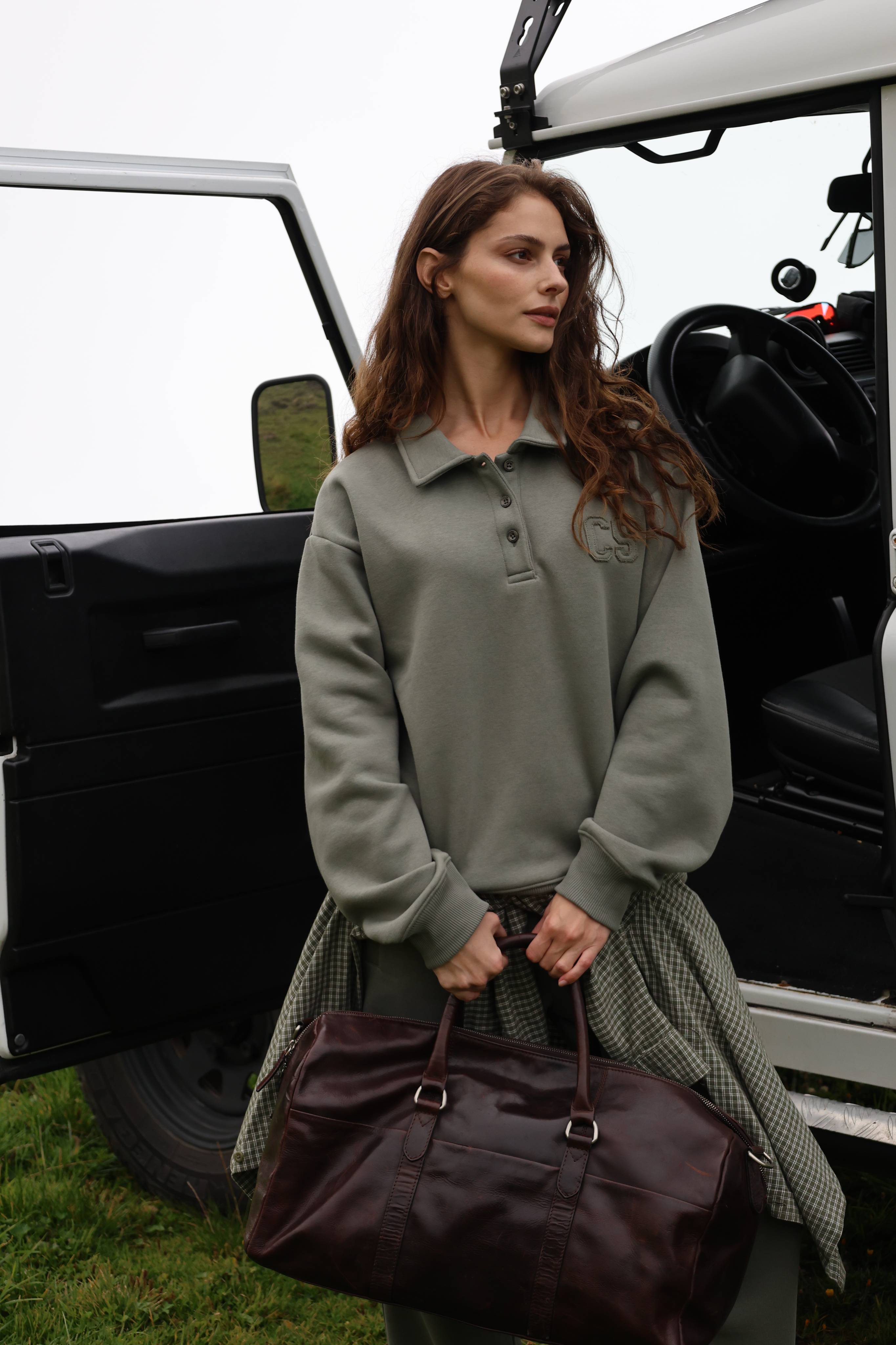 Woman wearing a khaki CASPARA CS rugby shirt holding a brown leather bag by a car.