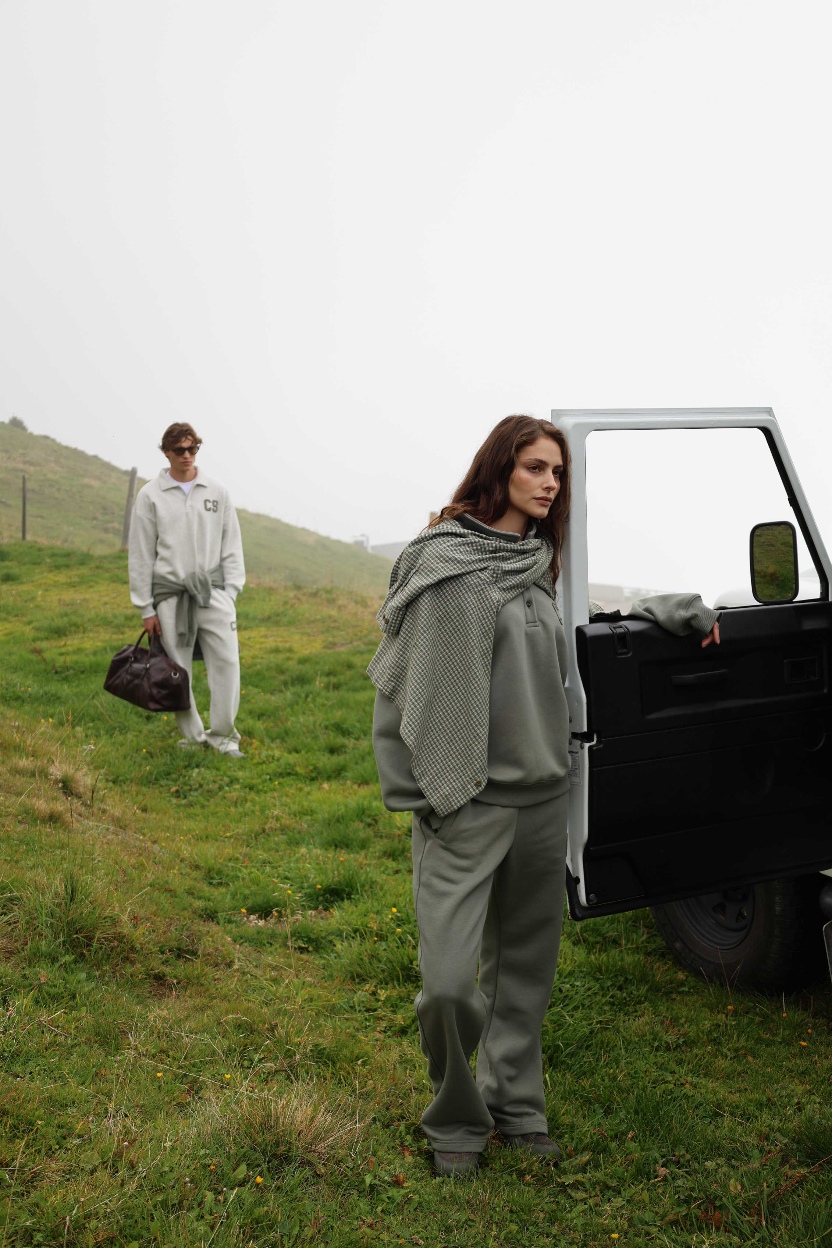 Woman wearing a khaki CASPARA CS rugby shirt and sweatpants standing outdoors beside a car.