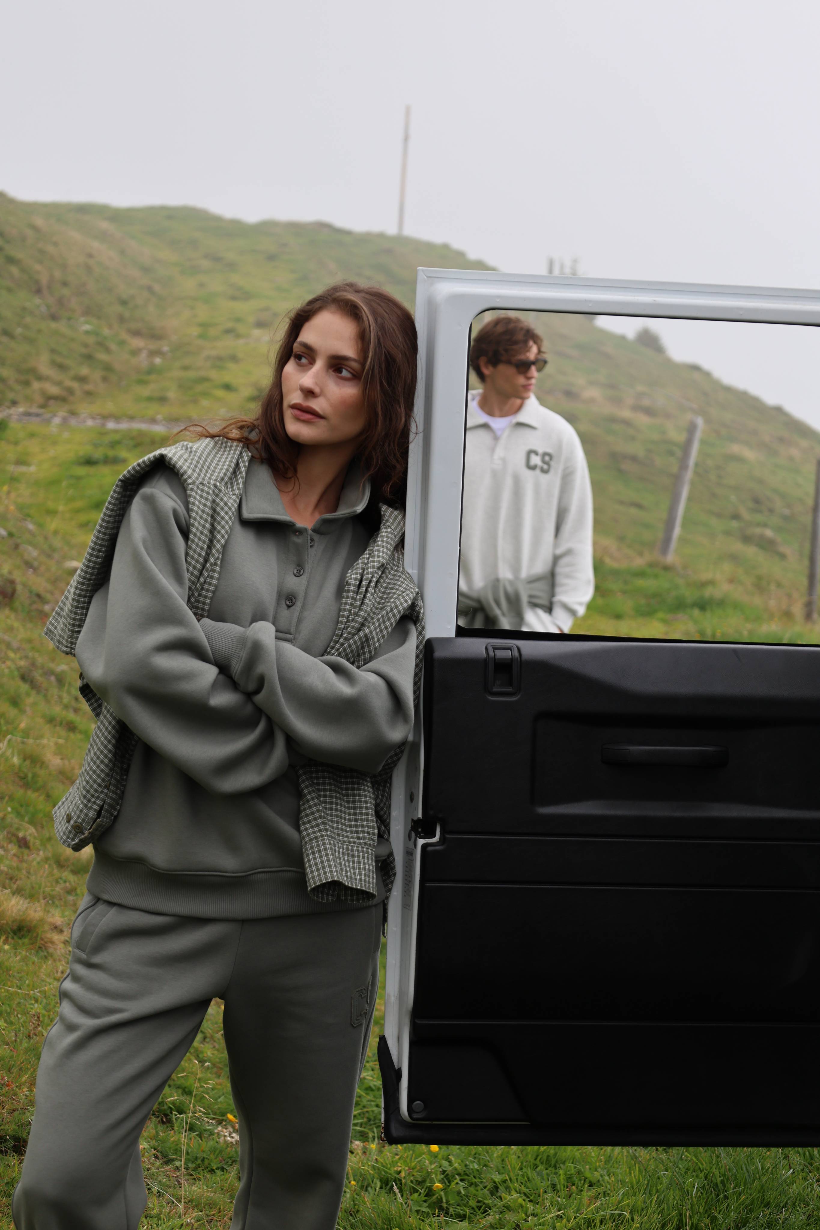 Woman wearing a khaki CASPARA CS rugby shirt and sweatpants leaning against a car outdoors.