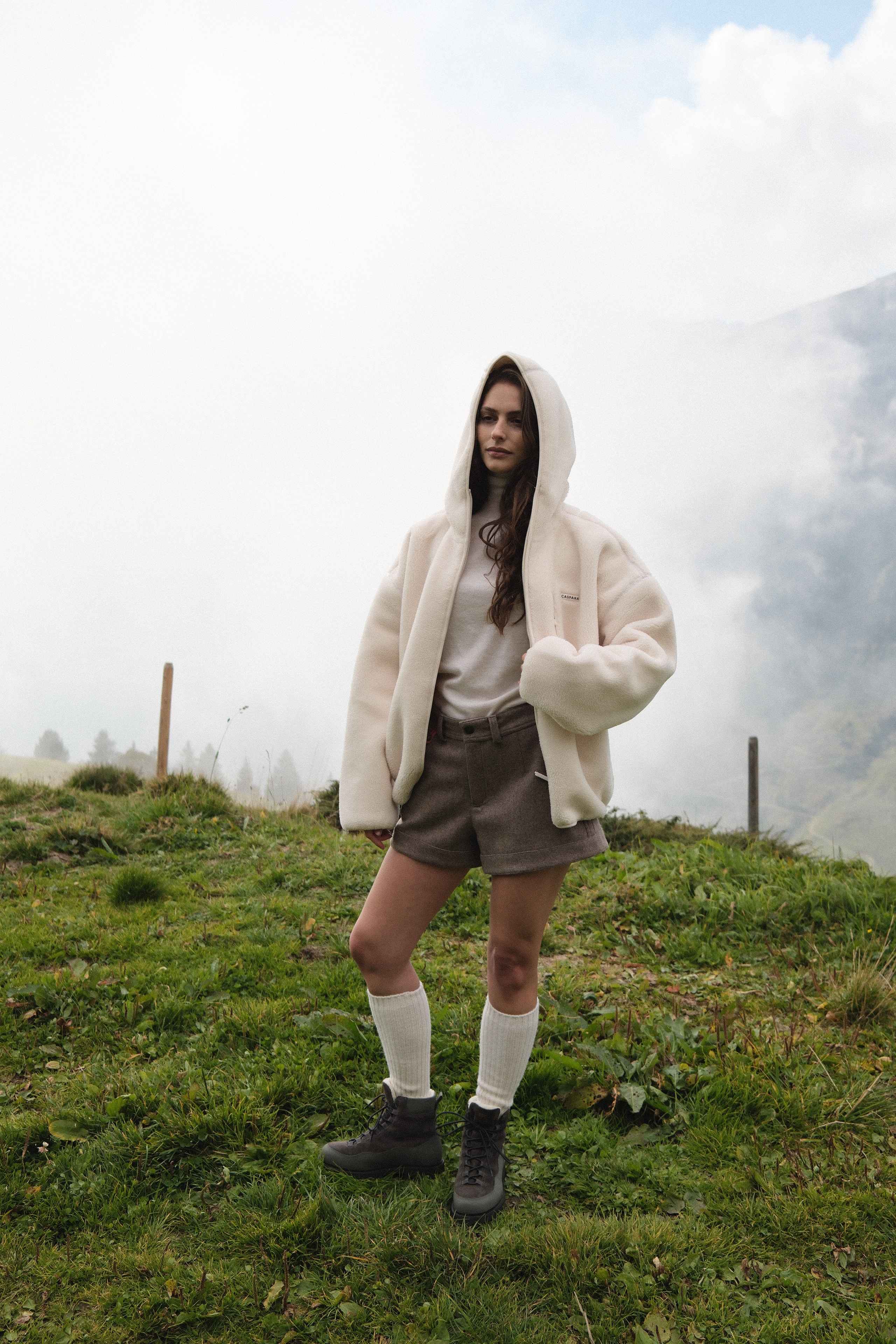 Woman wearing a creme CASPARA hooded fleece jacket outdoors on a grassy hillside.