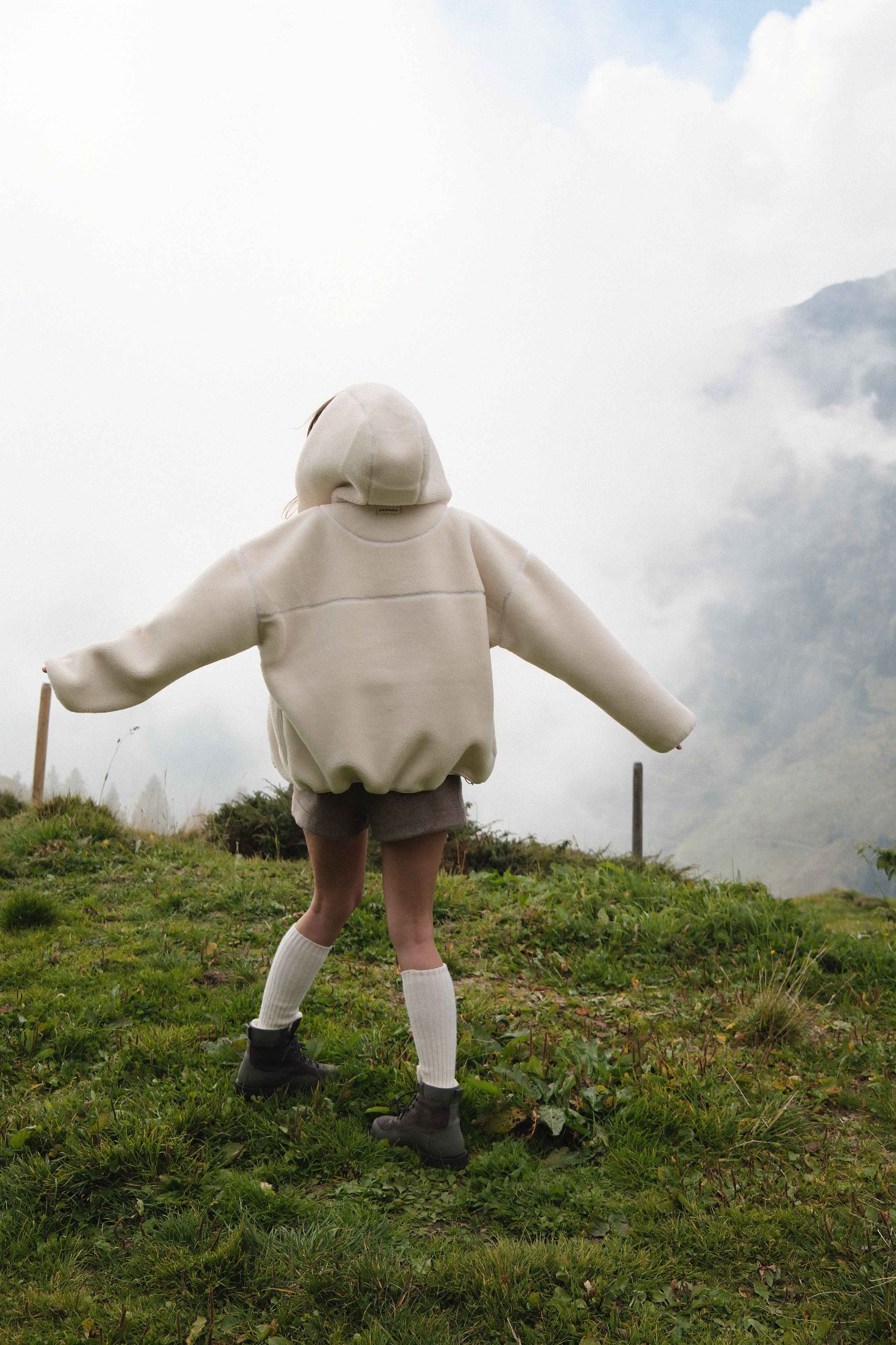 Back view of a woman wearing an oversized creme CASPARA Cloudfleece hooded jacket outdoors.
