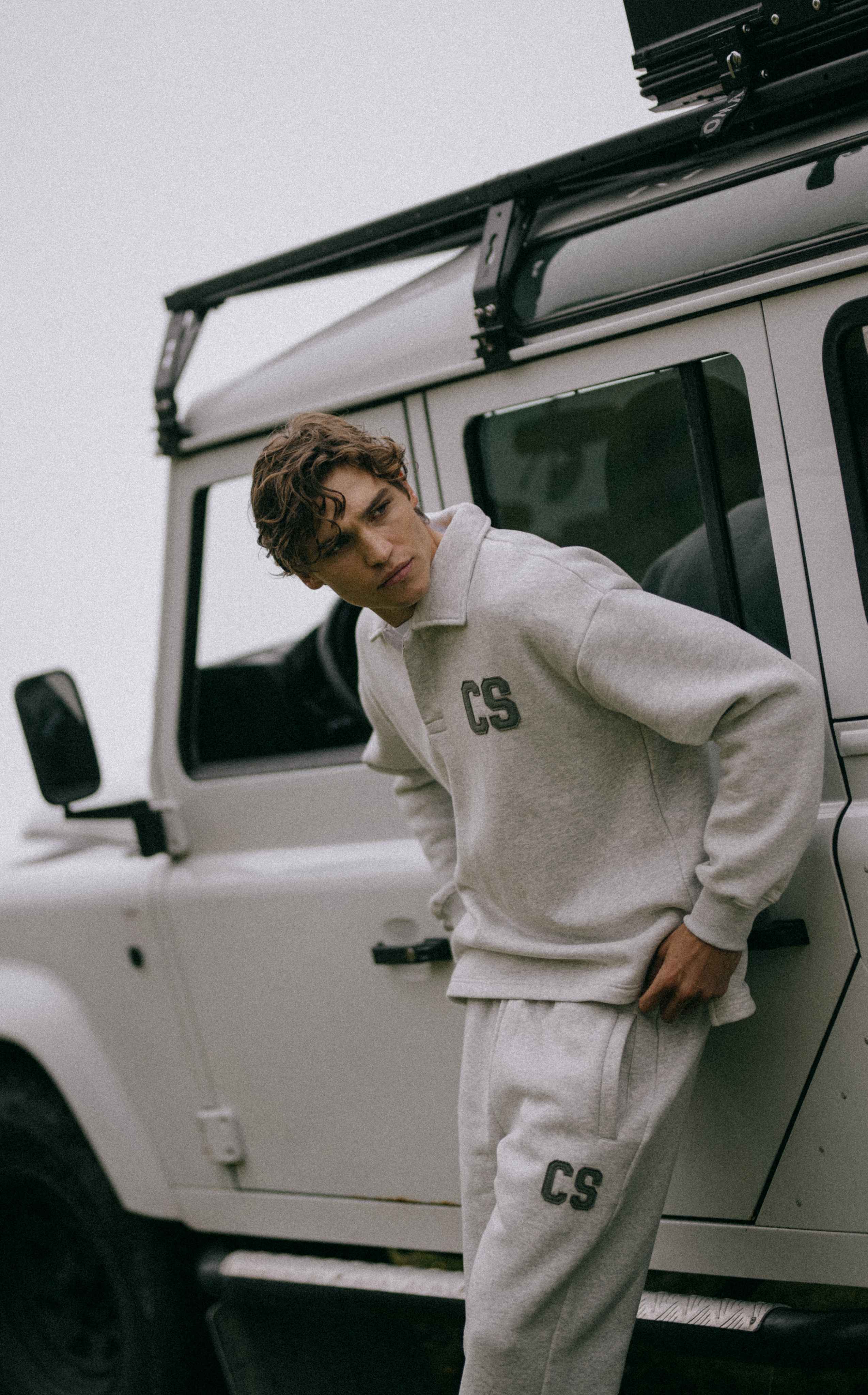 Man wearing a grey melange CASPARA CS rugby shirt and sweatpants leaning against an off-road vehicle.
