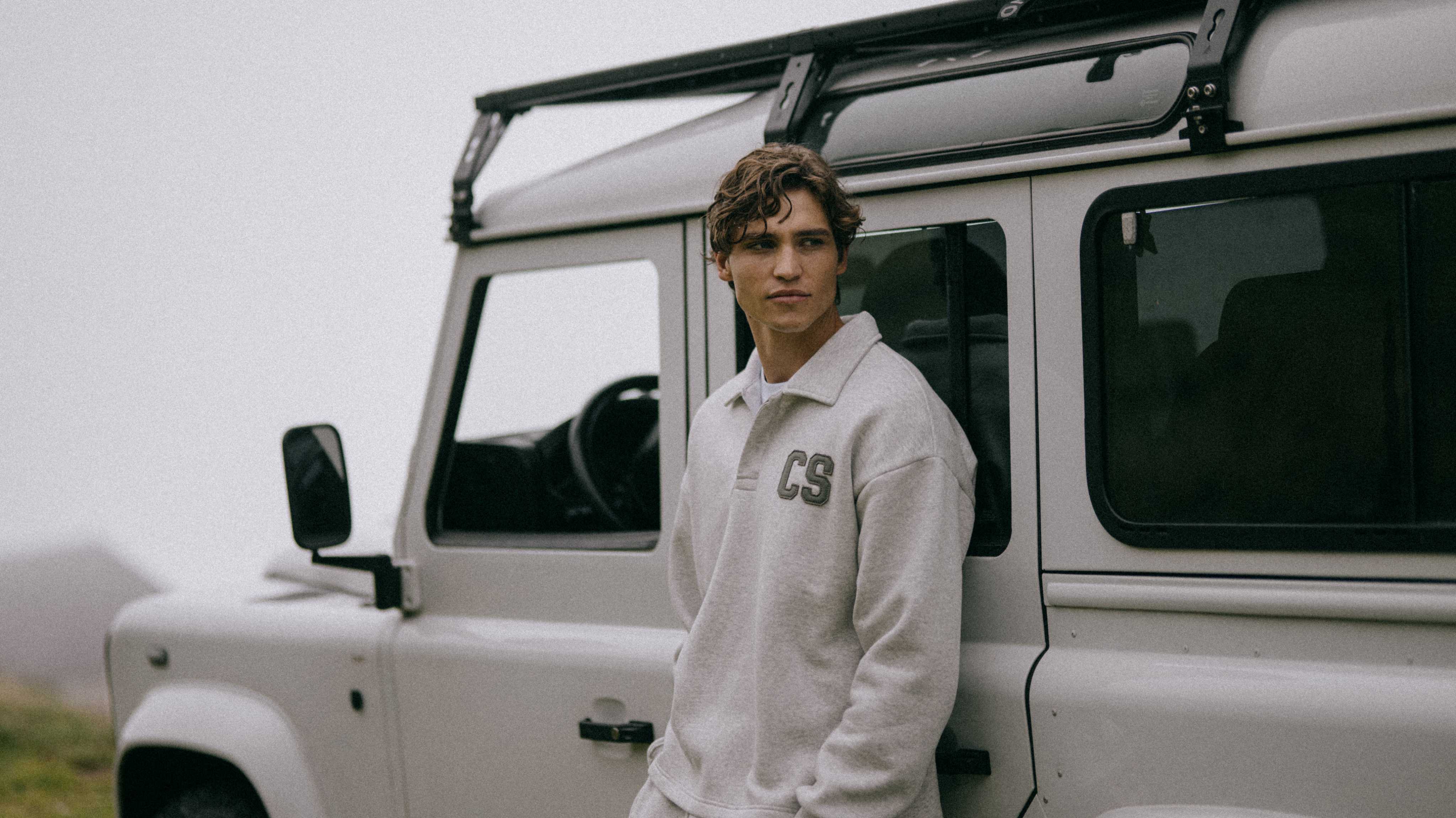 Man wearing a grey melange CASPARA CS rugby shirt leaning against an off-road vehicle.