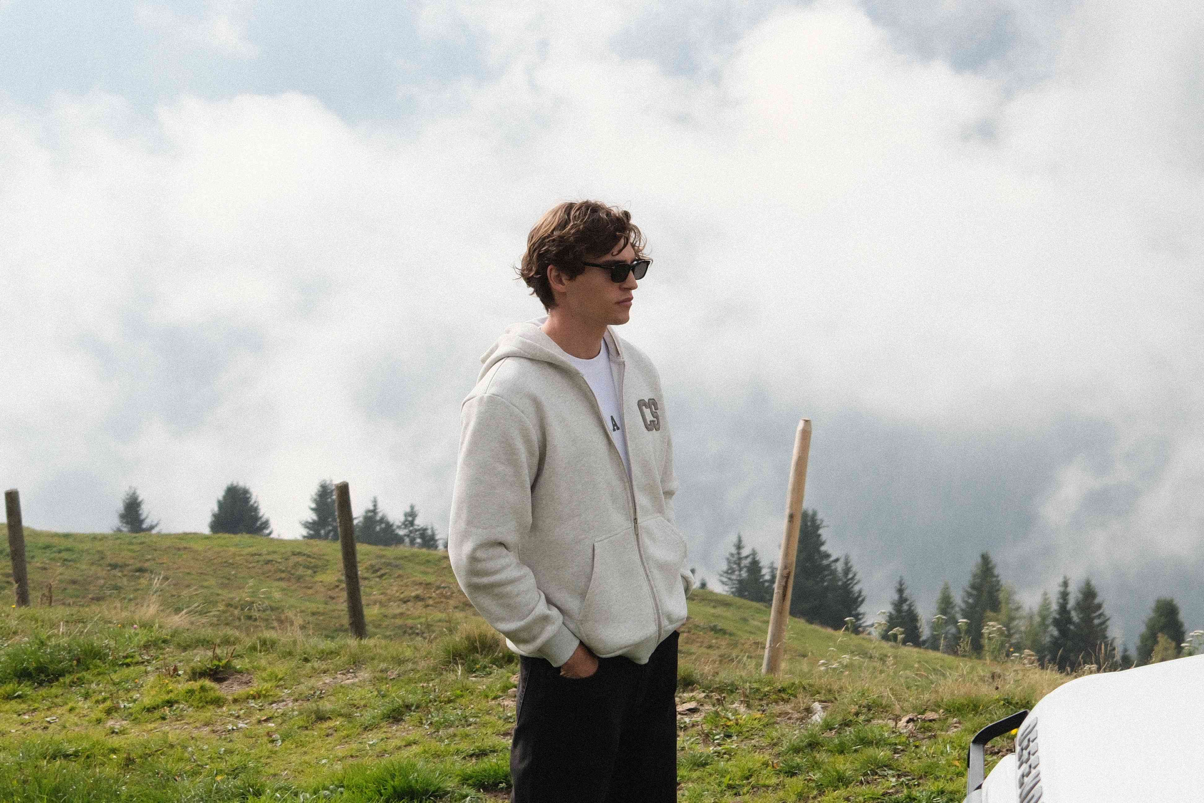 Man wearing a grey melange CASPARA CS zip hoodie standing on a grassy hillside.