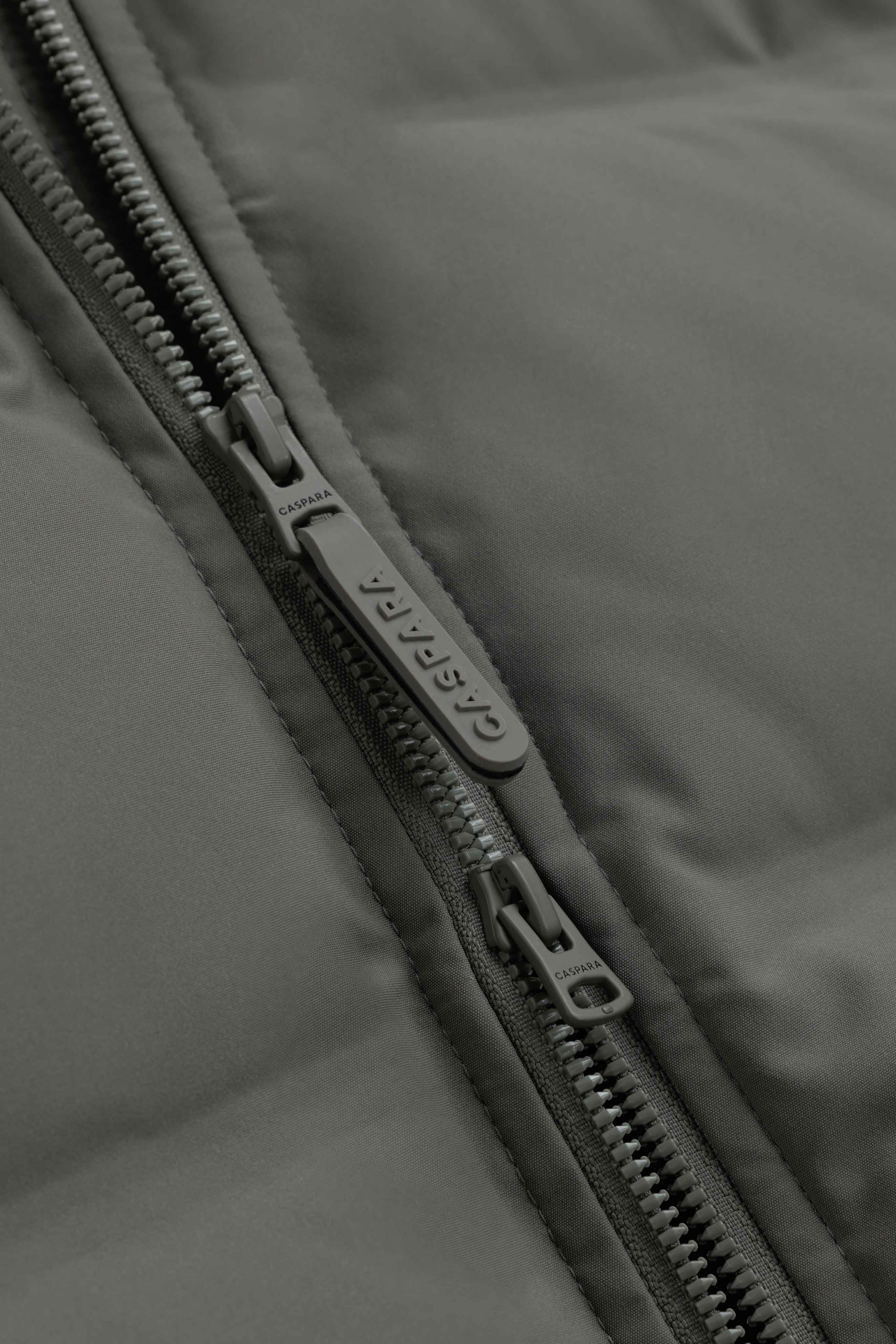Close-up of the front zipper and logo pull tab on the khaki CASPARA Aurora puffer jacket.