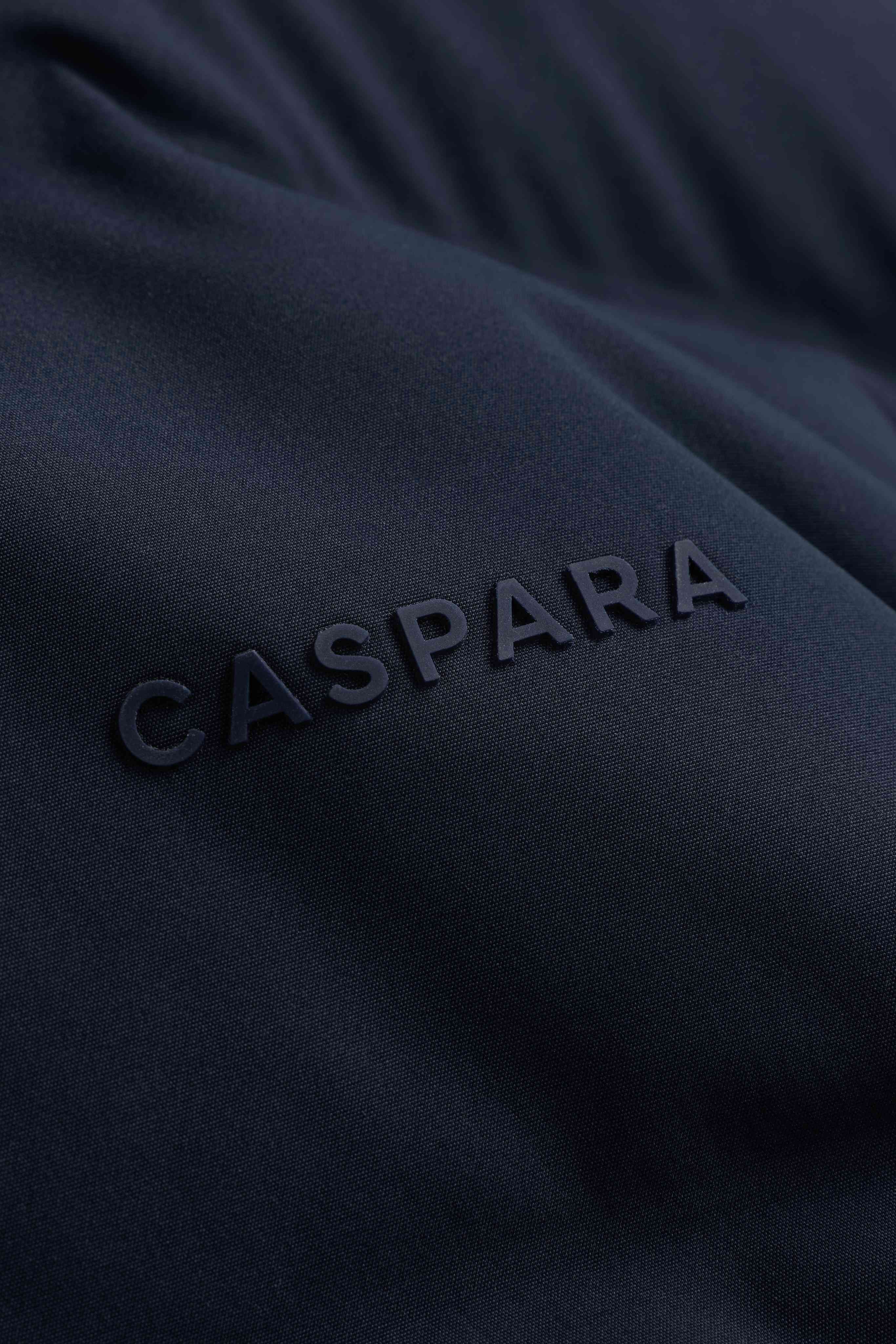 Close-up of the tonal CASPARA logo on the navy Aurora puffer jacket.