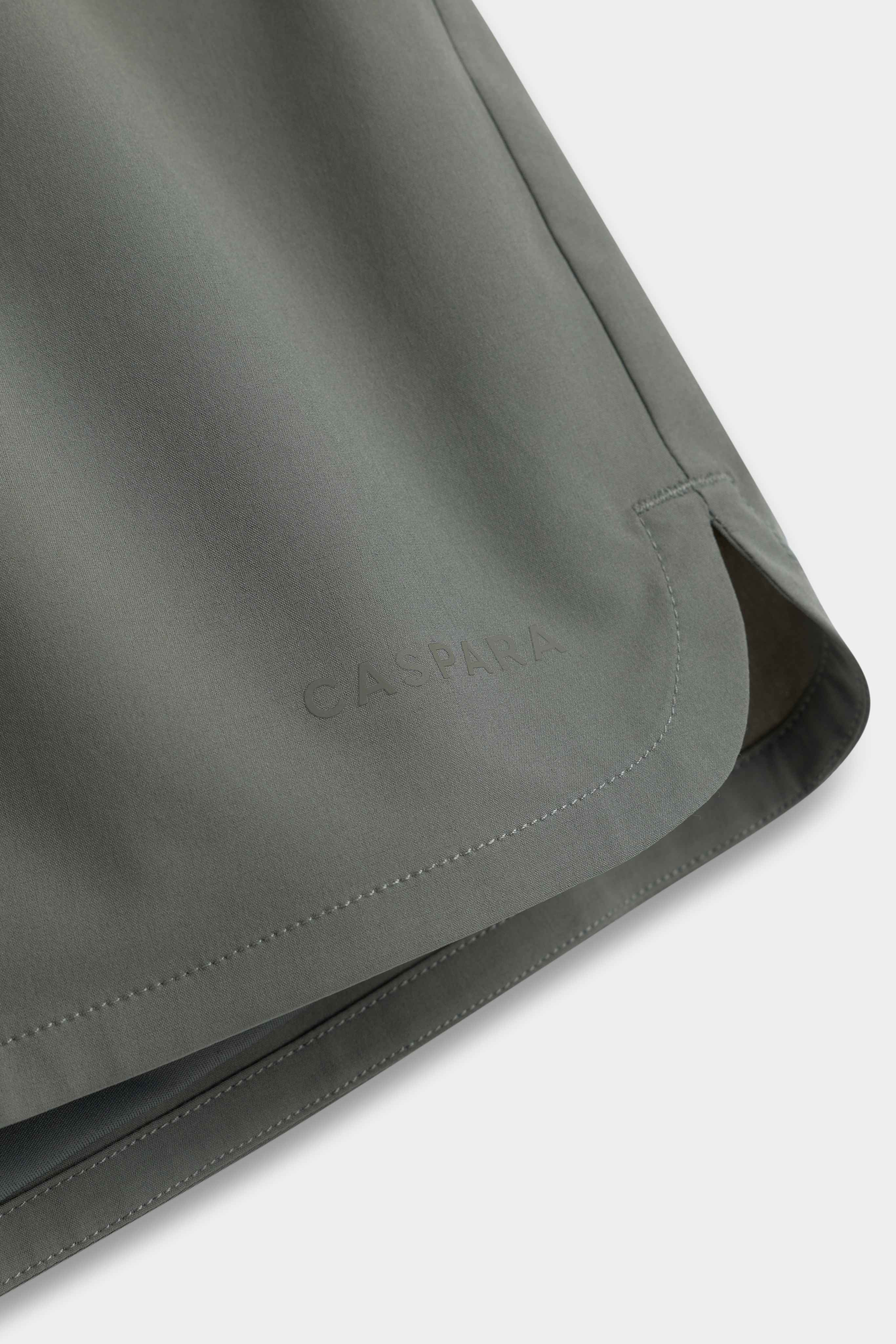 Close-up of the hem on khaki CASPARA Aerluxe shorts featuring a subtle tonal logo and side slit.