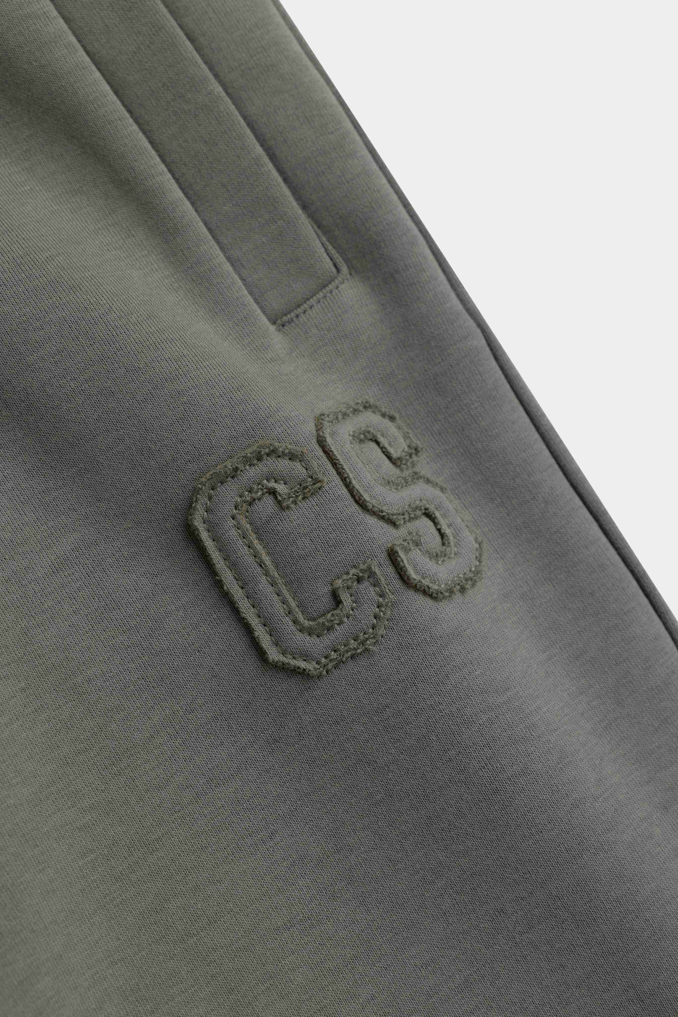 Close-up of khaki CASPARA sweatpants showing an embroidered CS logo and side pocket detail.