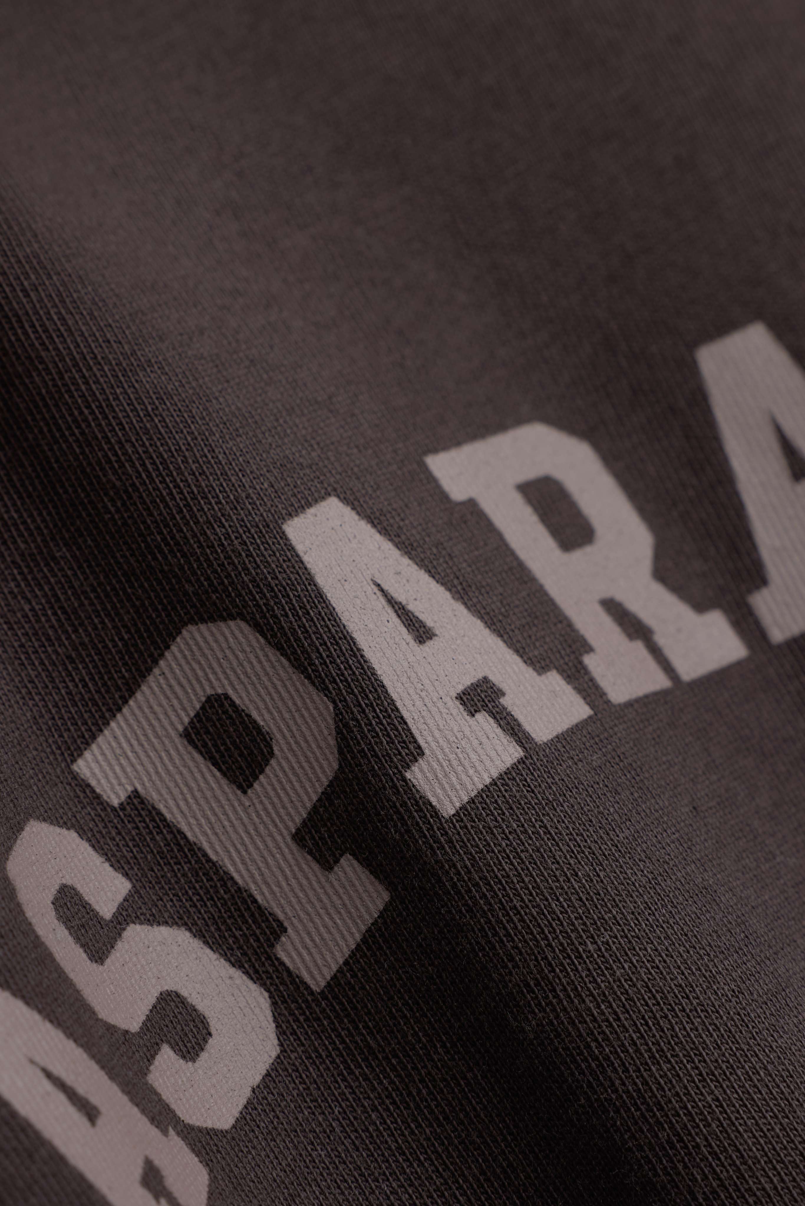 Close-up of the light brown CASPARA logo printed on the espresso CS Signature long sleeve.