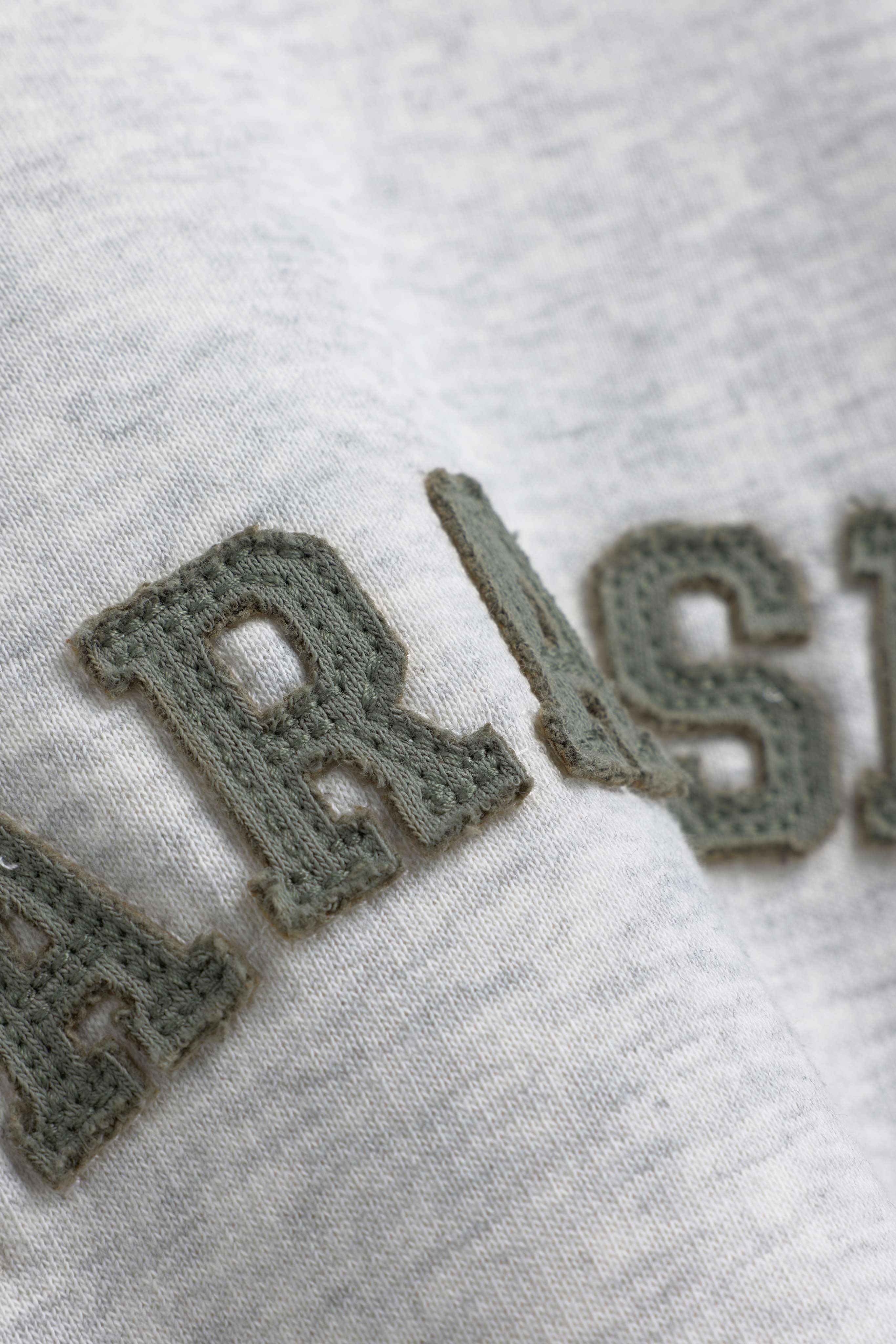 Close-up of khaki embroidered lettering on the grey melange CASPARA CS crewneck sweatshirt.