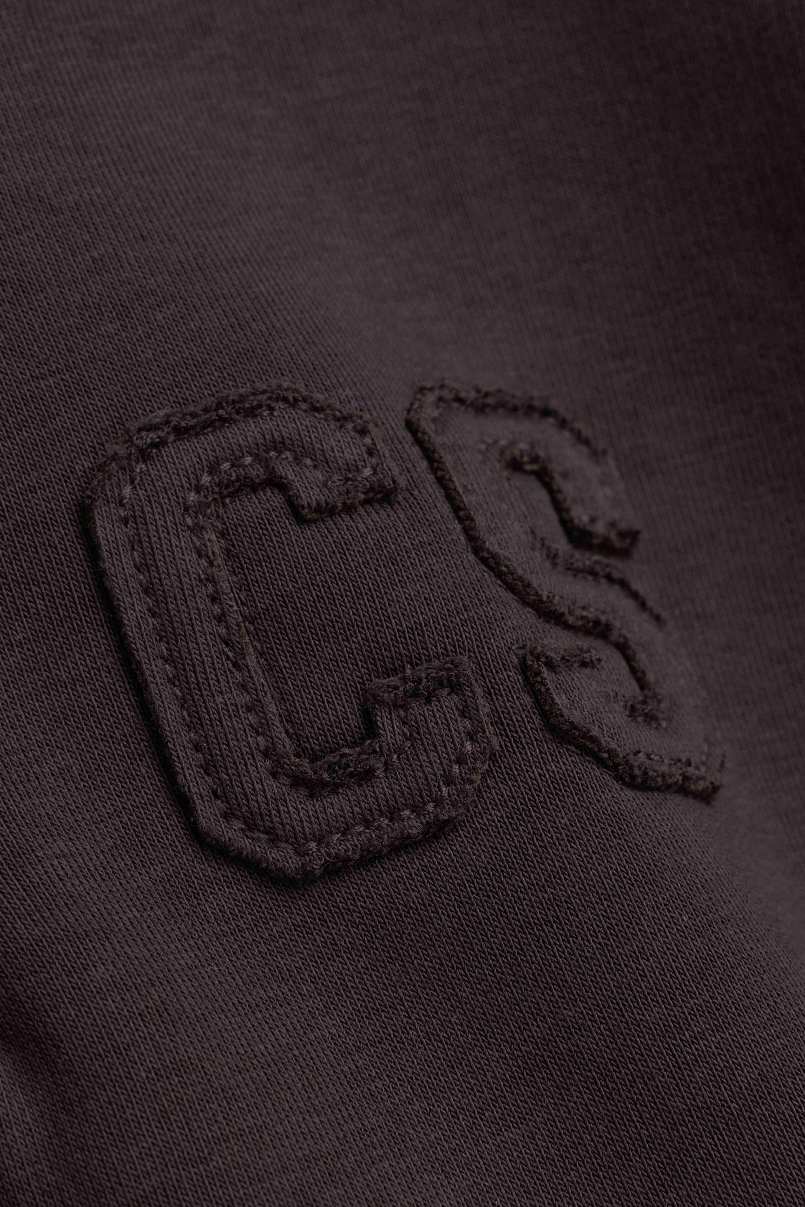 Close-up of the tonal embroidered CS logo on an espresso CASPARA rugby shirt.