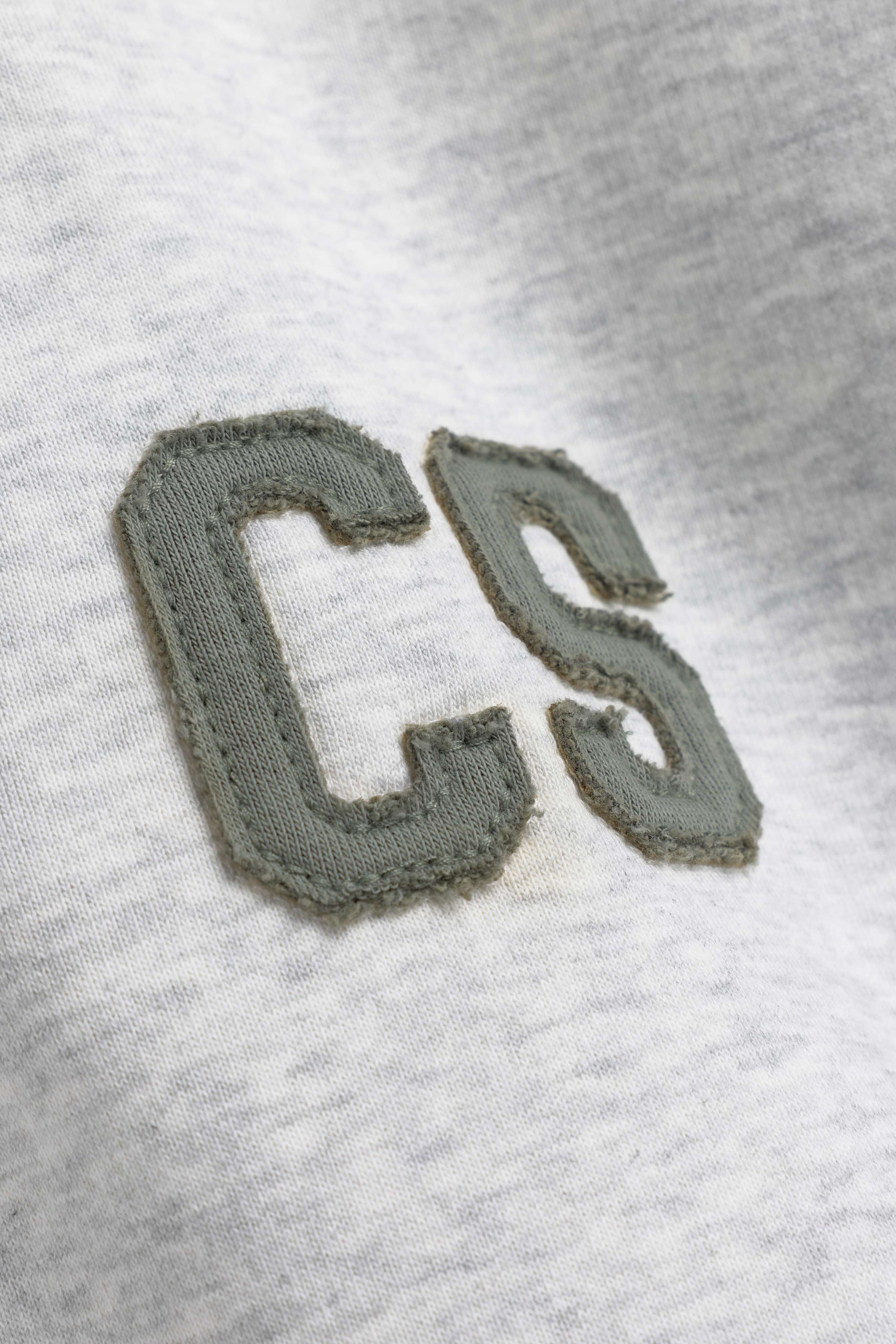 Close-up of the khaki embroidered CS logo patch on a grey melange CASPARA rugby shirt.
