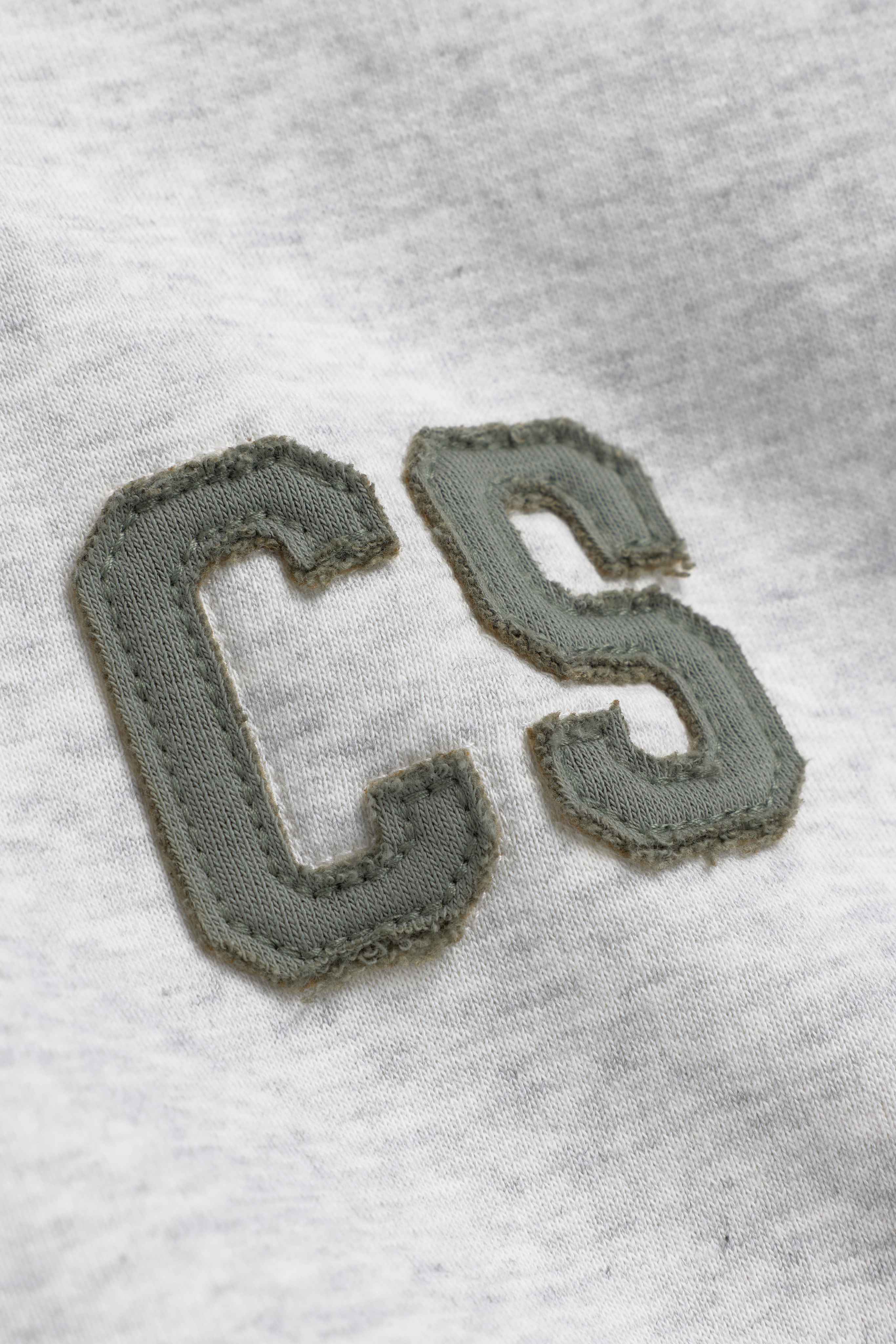 Close-up of khaki CS patch detail on a grey melange CASPARA hoodie.