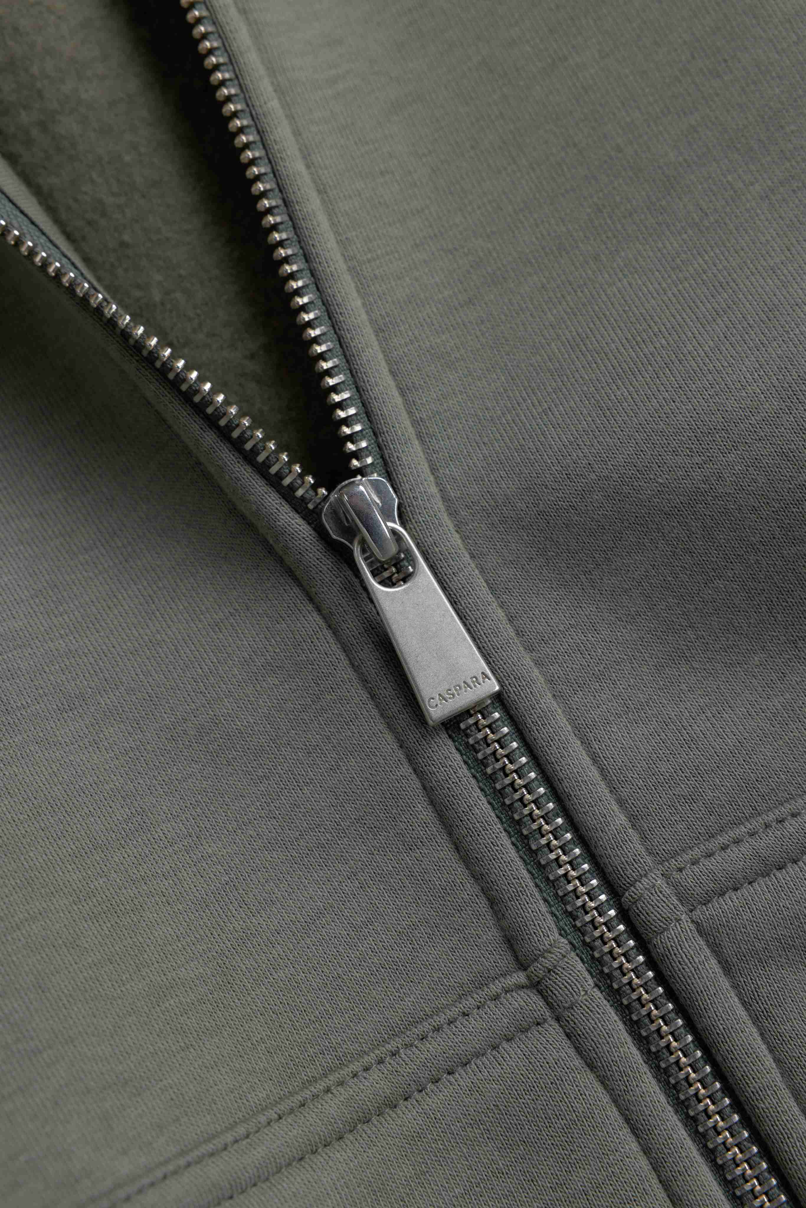Close-up of the engraved silver zipper pull on a khaki CASPARA CS Zip Hoodie.