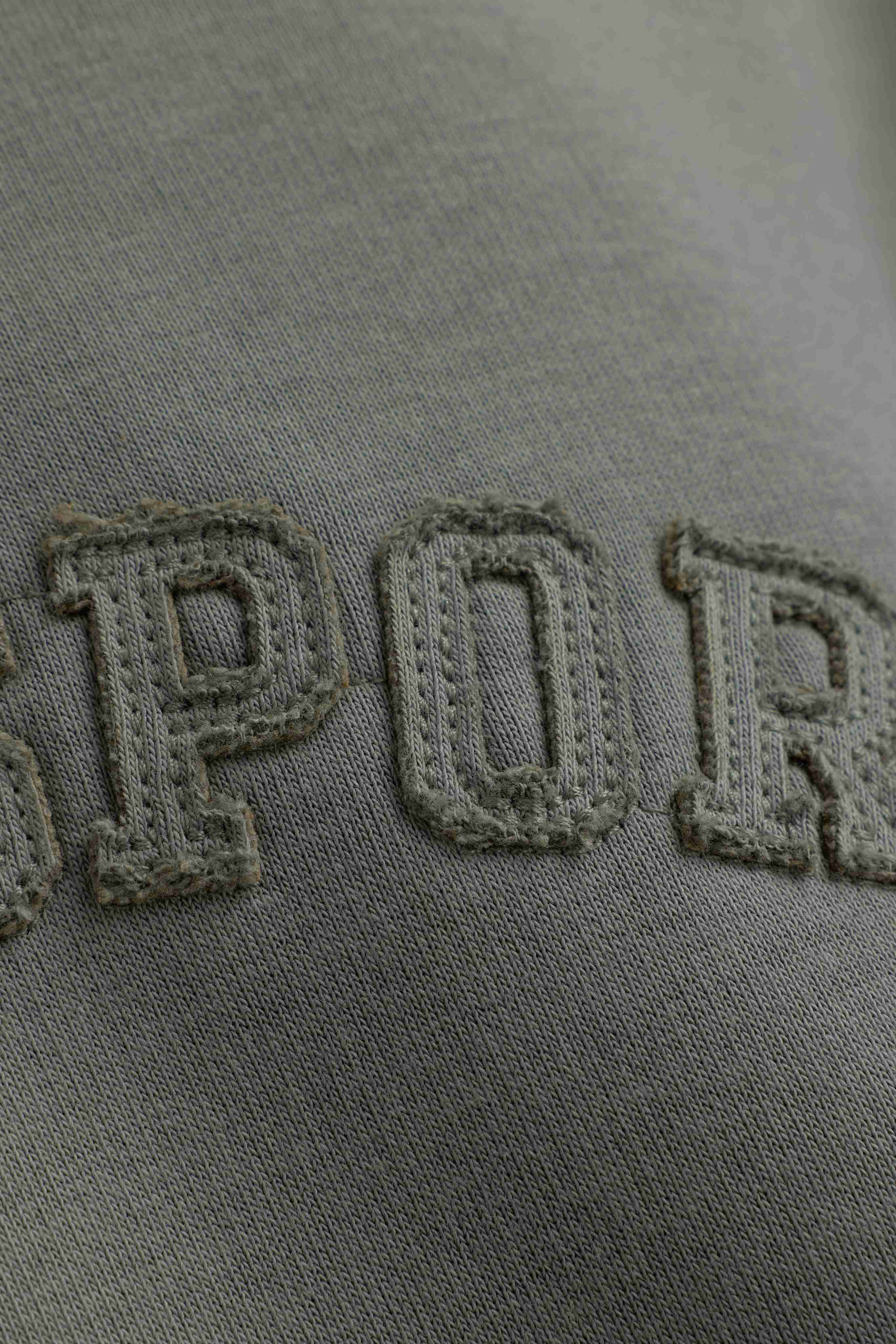 Close-up of stitched appliqué lettering on the fabric of a khaki CASPARA CS hoodie.