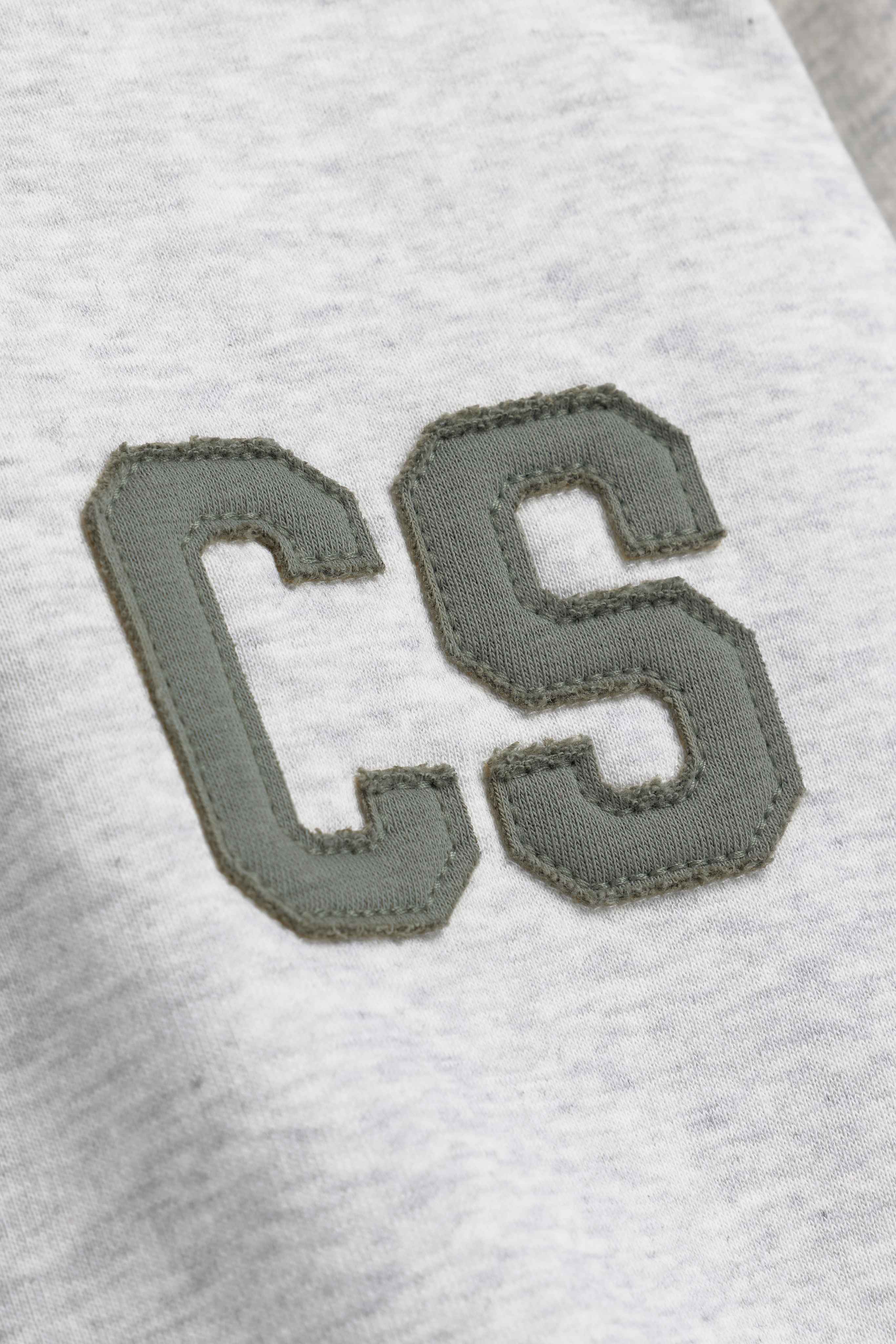Close-up of the khaki CS patch stitched on a grey melange CASPARA hoodie.