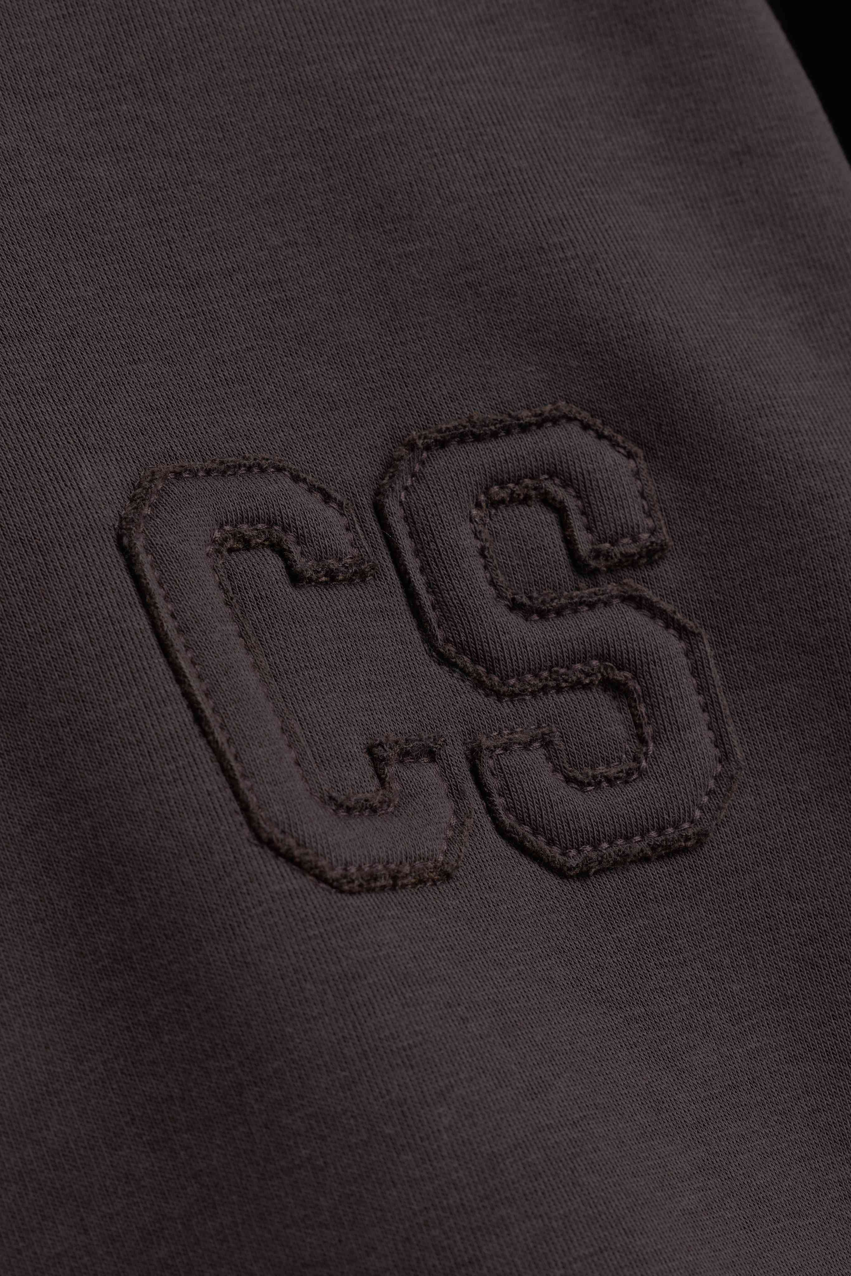 Close-up of the tonal embroidered CS logo on the espresso CASPARA CS Zip Hoodie.
