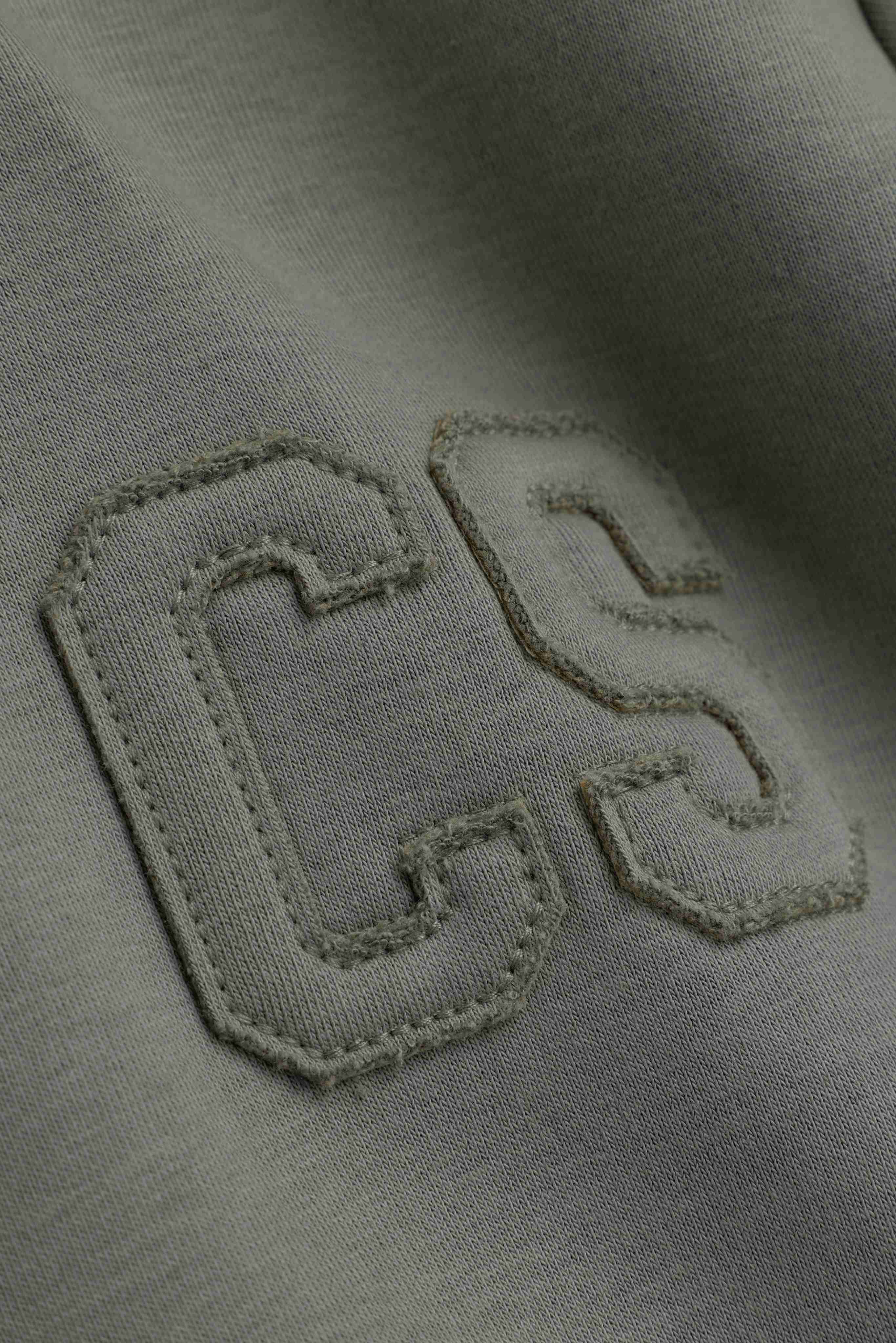 Close-up of the stitched CS logo detail on a khaki CASPARA zip hoodie.