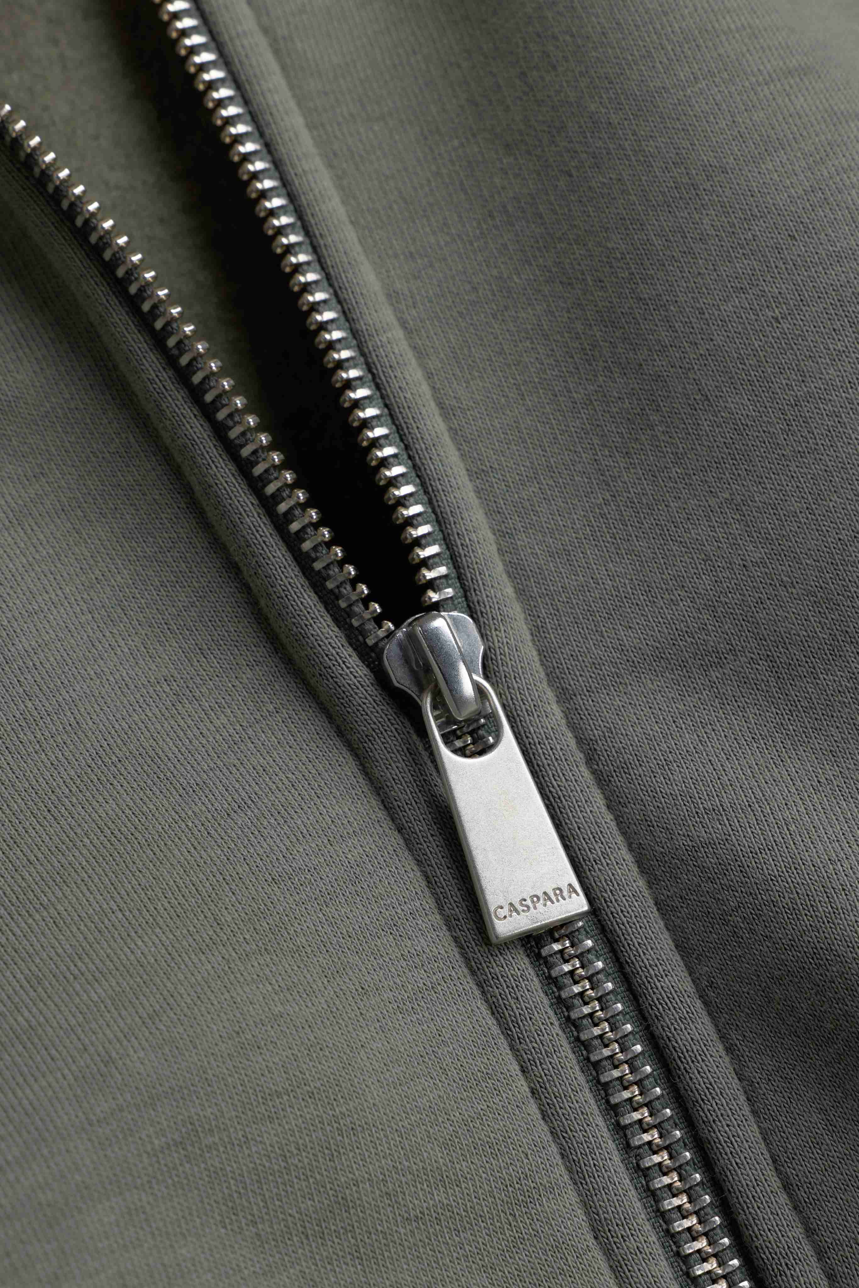 Close-up of the silver zipper pull engraved with CASPARA on a khaki CS Zip Hoodie.