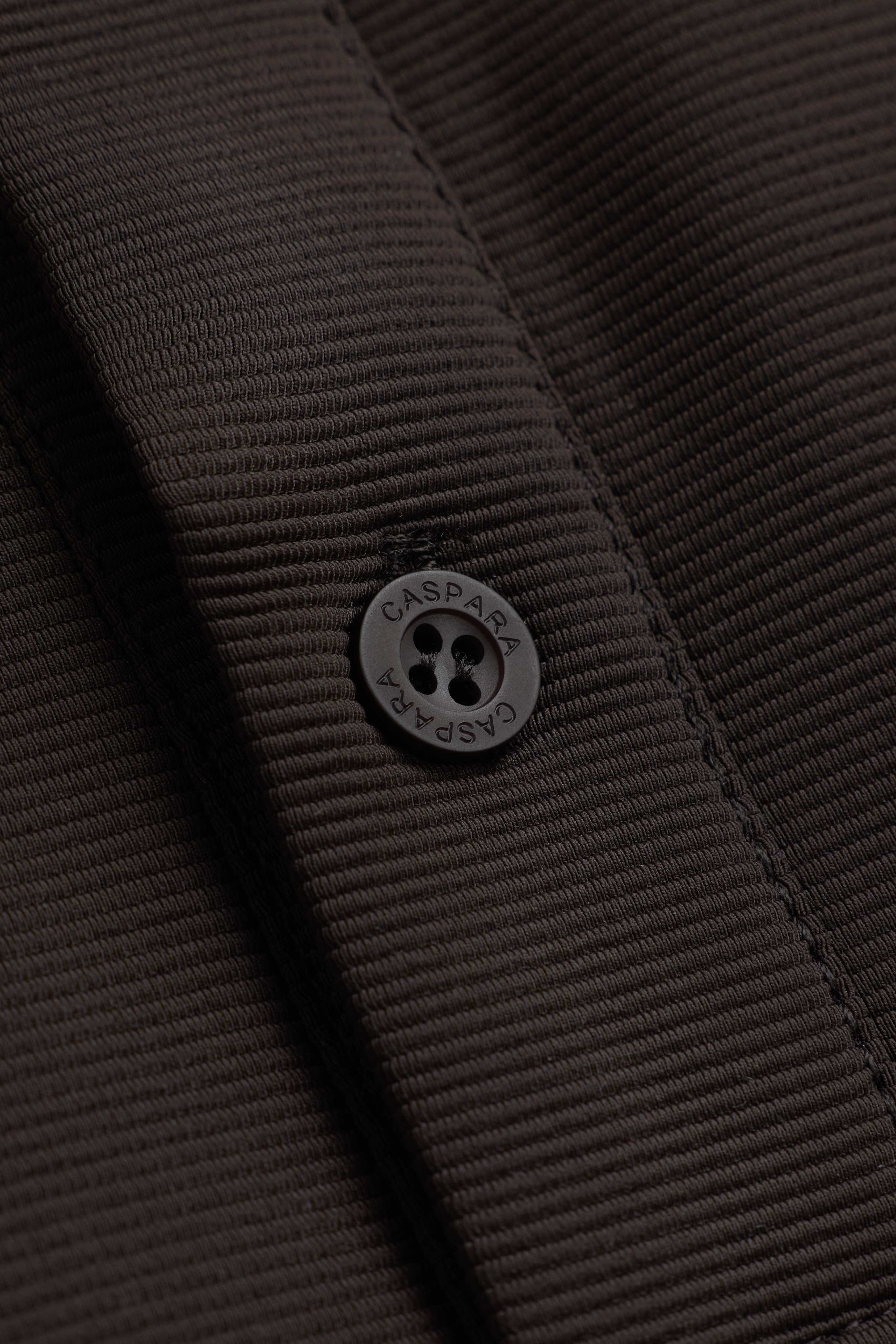 Close-up of the engraved button and textured fabric on the espresso CASPARA Vantage polo tee.
