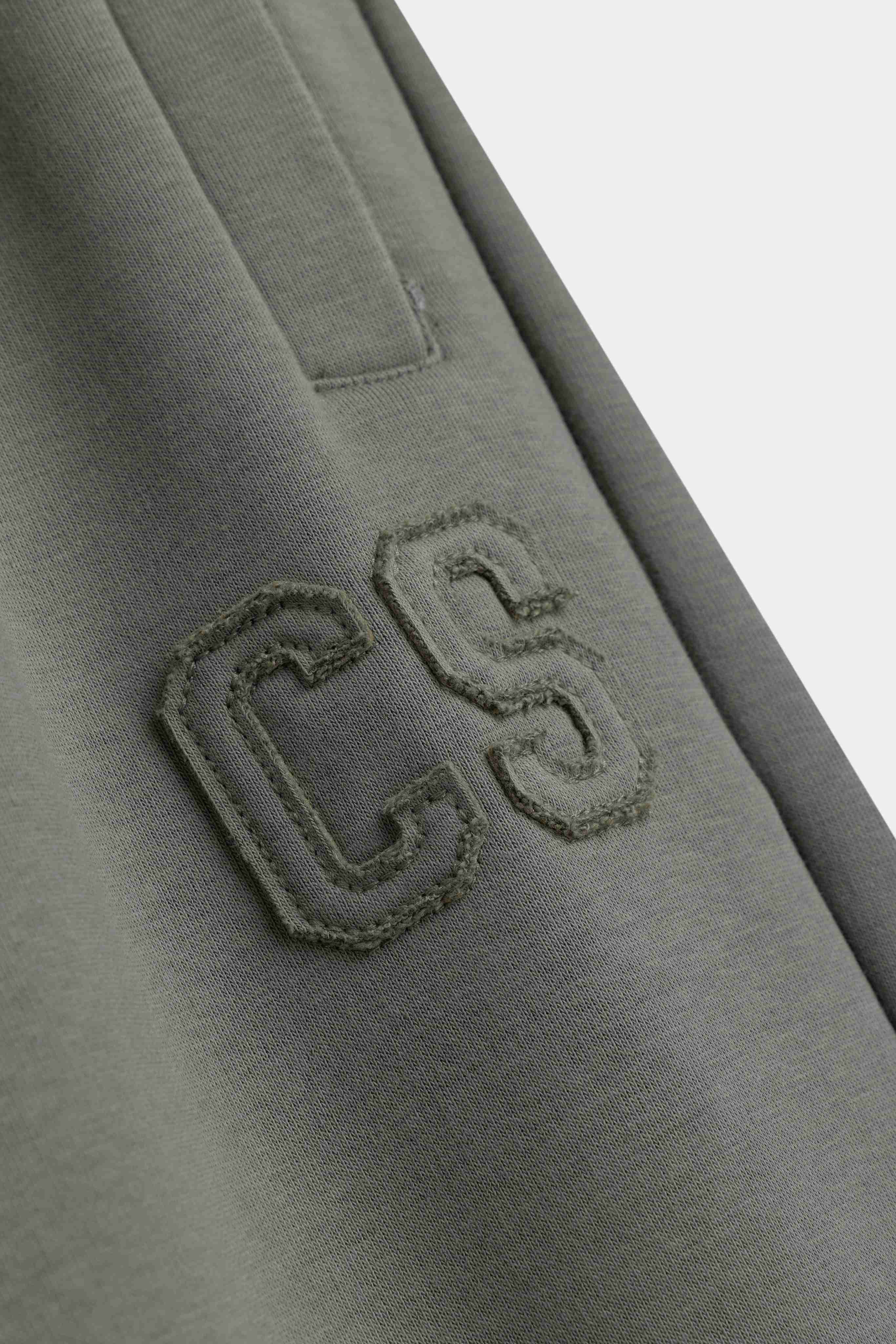 Close-up of the embroidered CS logo and pocket detail on khaki CASPARA sweatpants.