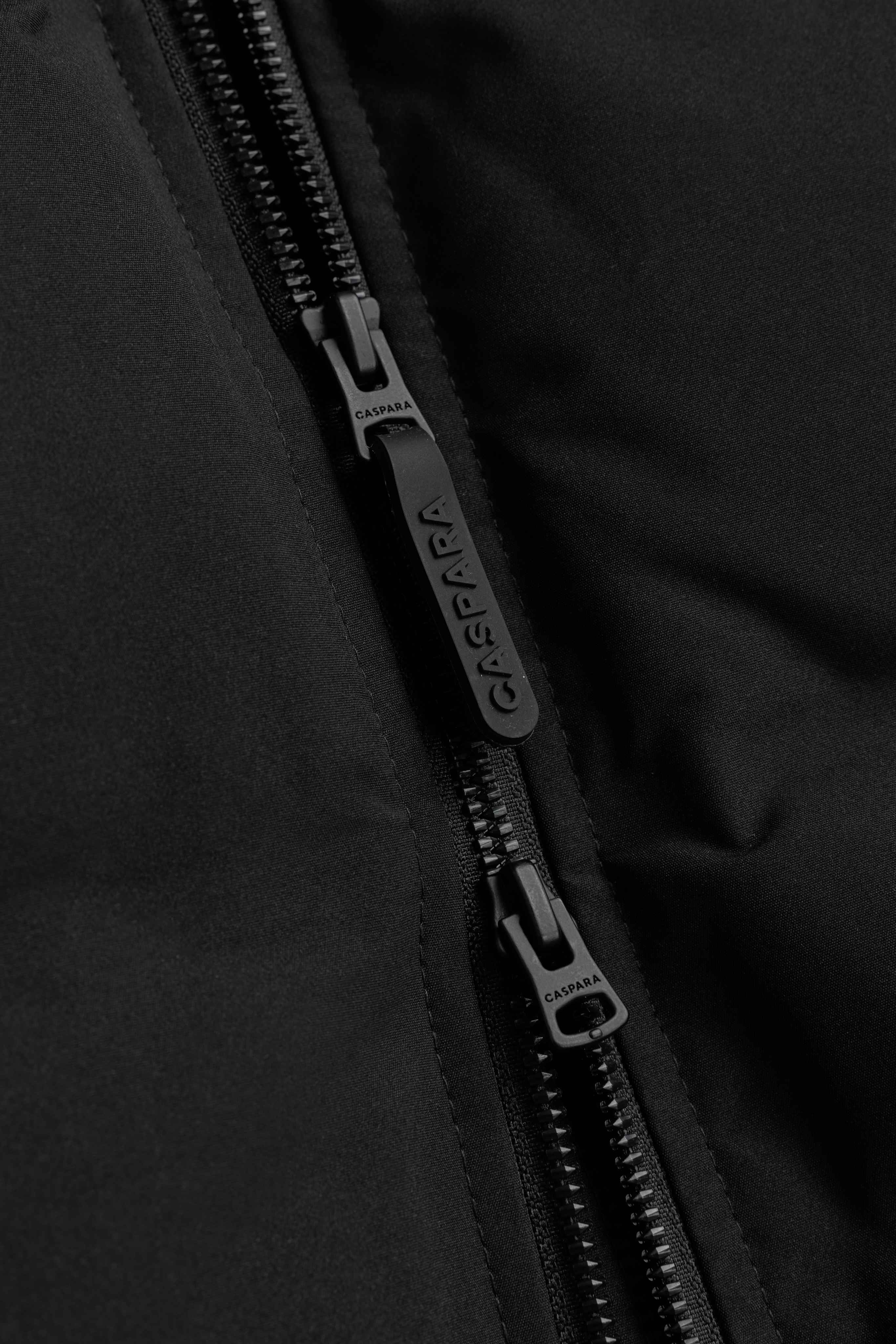 Close-up of the two-way front zipper with a branded pull tab on the black CASPARA Aurora puffer jacket.