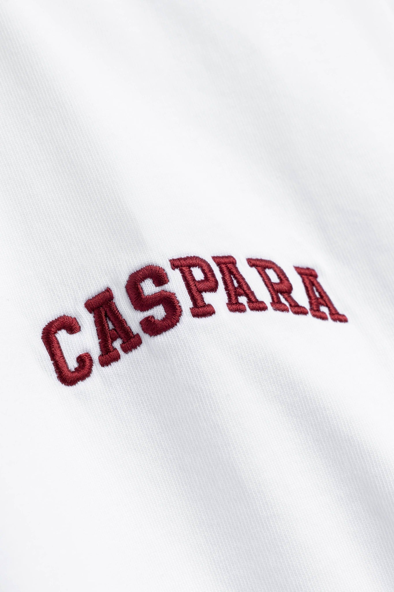 Close-up of burgundy embroidered CASPARA lettering on a white organic cotton varsity tee.