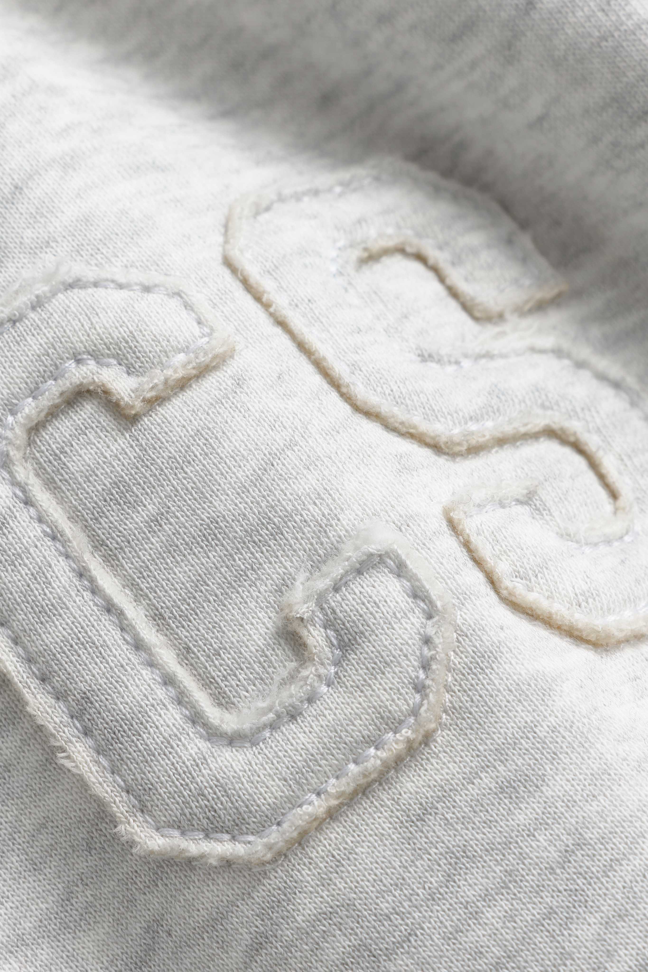 Close-up of the stitched CS logo patch on a grey melange CASPARA zip hoodie.