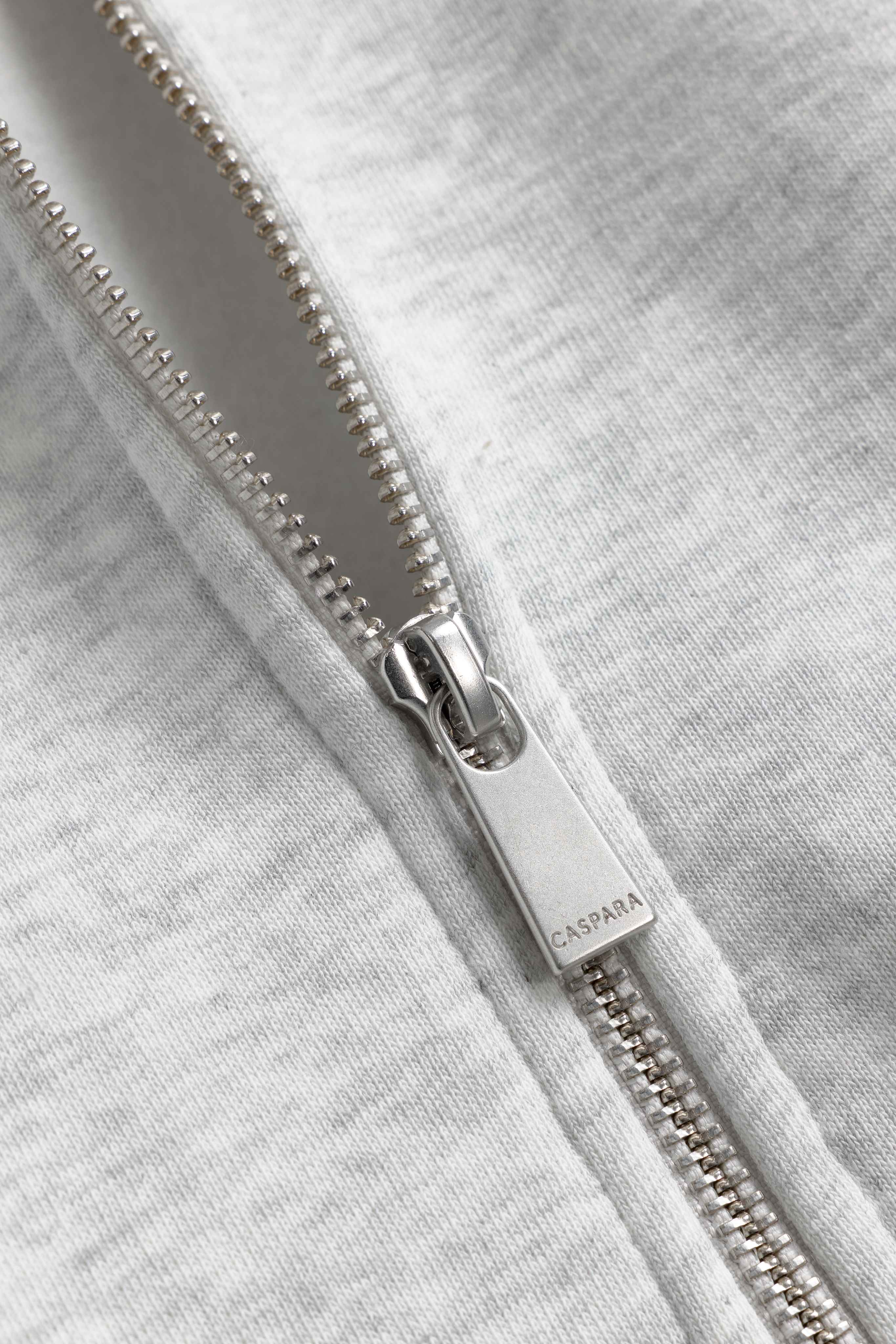 Close-up of the silver metal zipper pull engraved with the CASPARA logo on a grey melange zip hoodie.