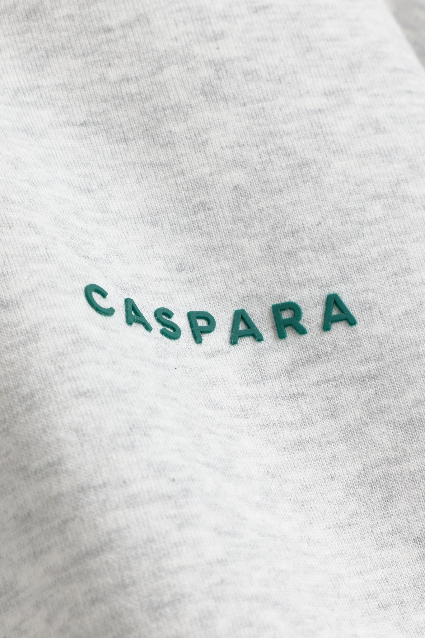 Close-up of the embossed green CASPARA logo on a grey melange sweatshirt.