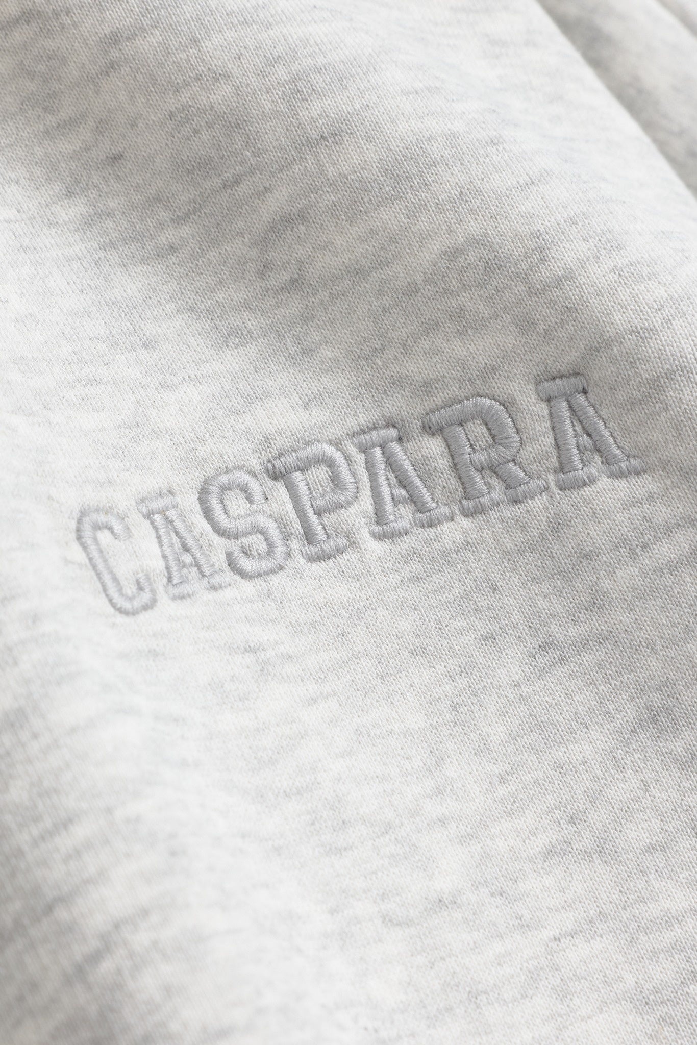 Close-up of embroidered CASPARA logo on grey melange sweatshirt fabric.