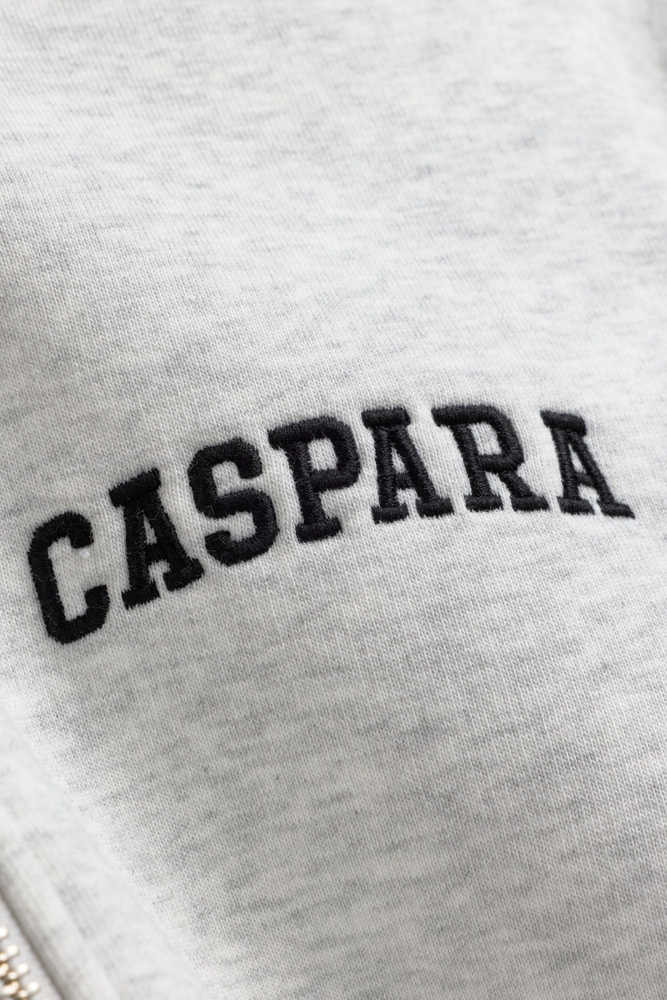 Close-up of black embroidered CASPARA logo on a grey melange zip hoodie.