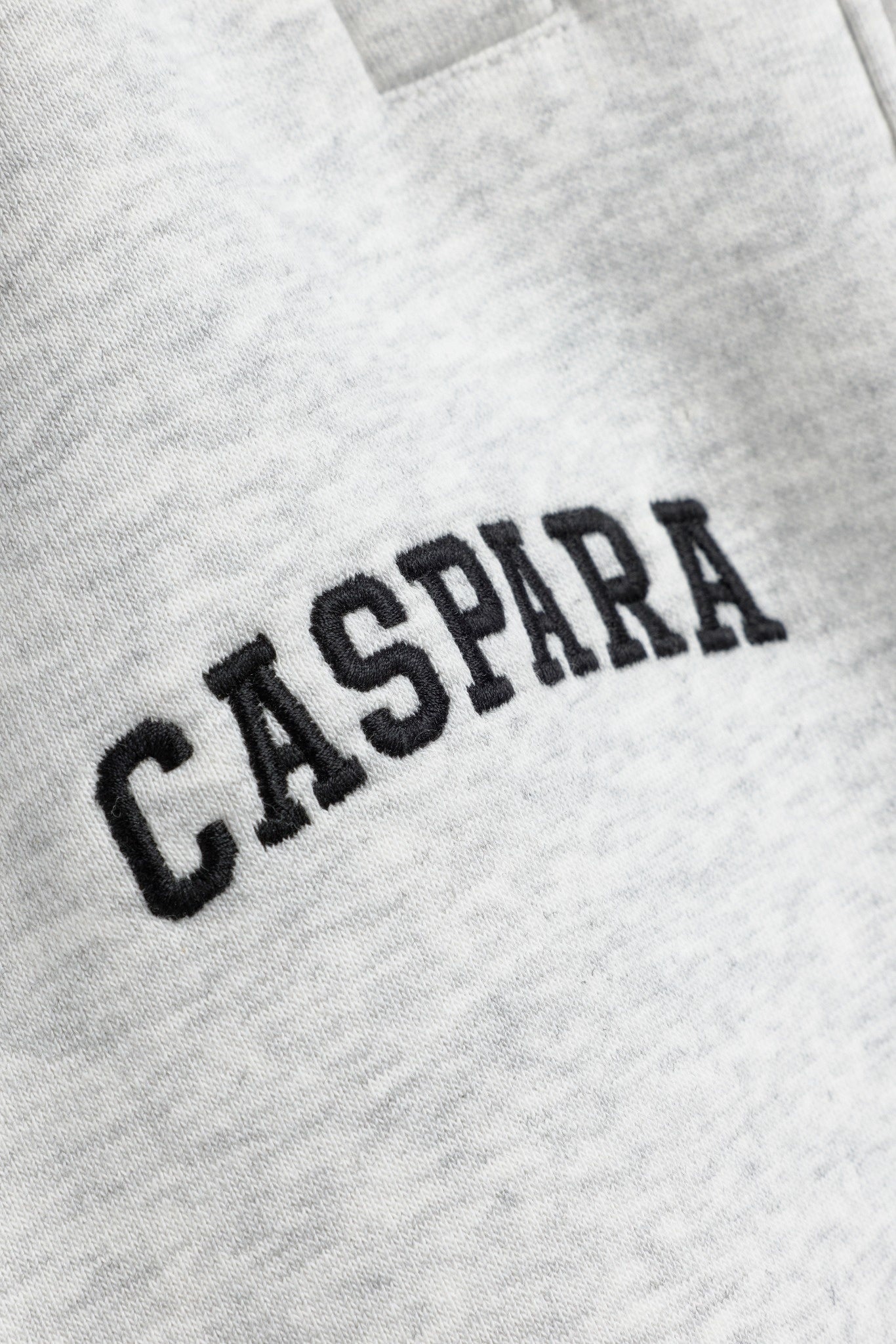 Close-up of black embroidered CASPARA logo on grey melange sweatpants.