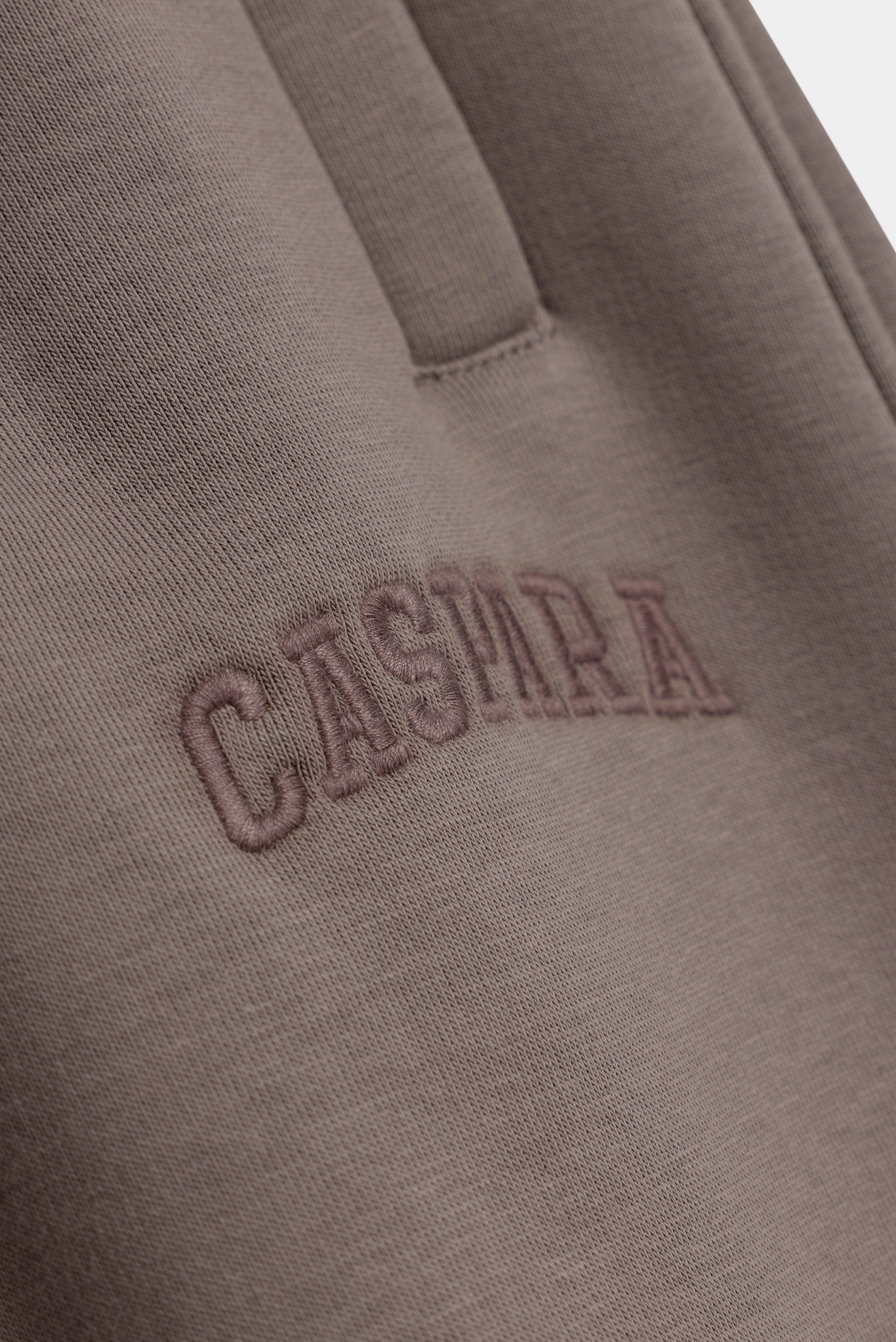 Close-up of embroidered CASPARA logo on walnut brown sweatpants.