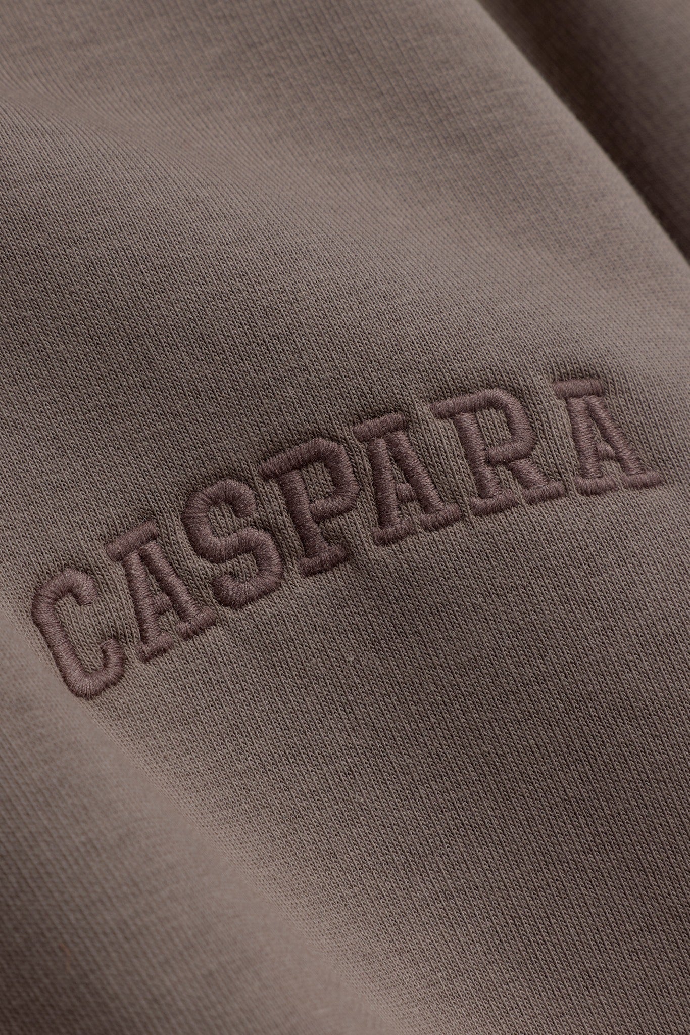 Close-up of tonal embroidered CASPARA logo on walnut brown hoodie fabric.