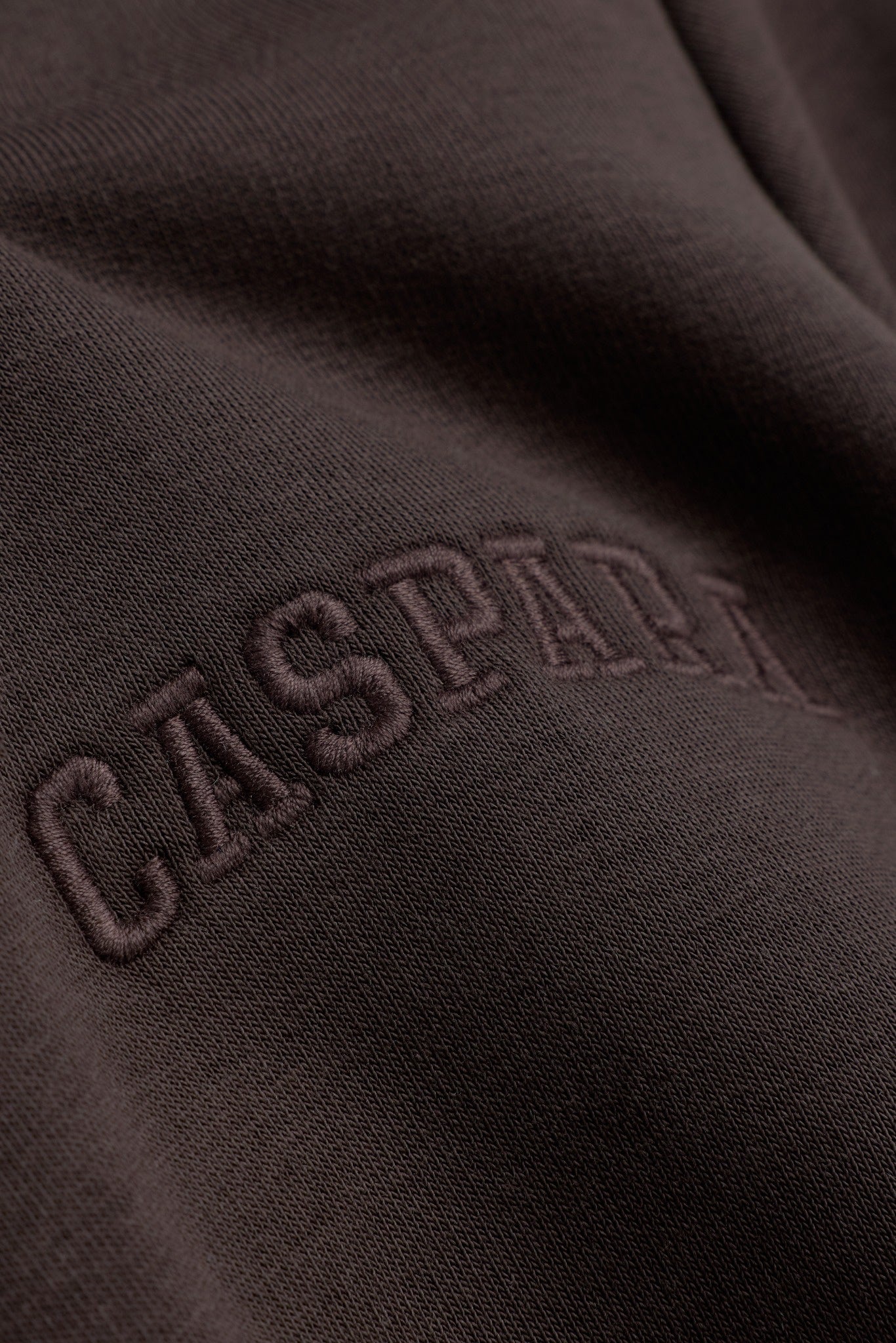 Close-up of embroidered CASPARA logo lettering on espresso brown cotton hoodie fabric.