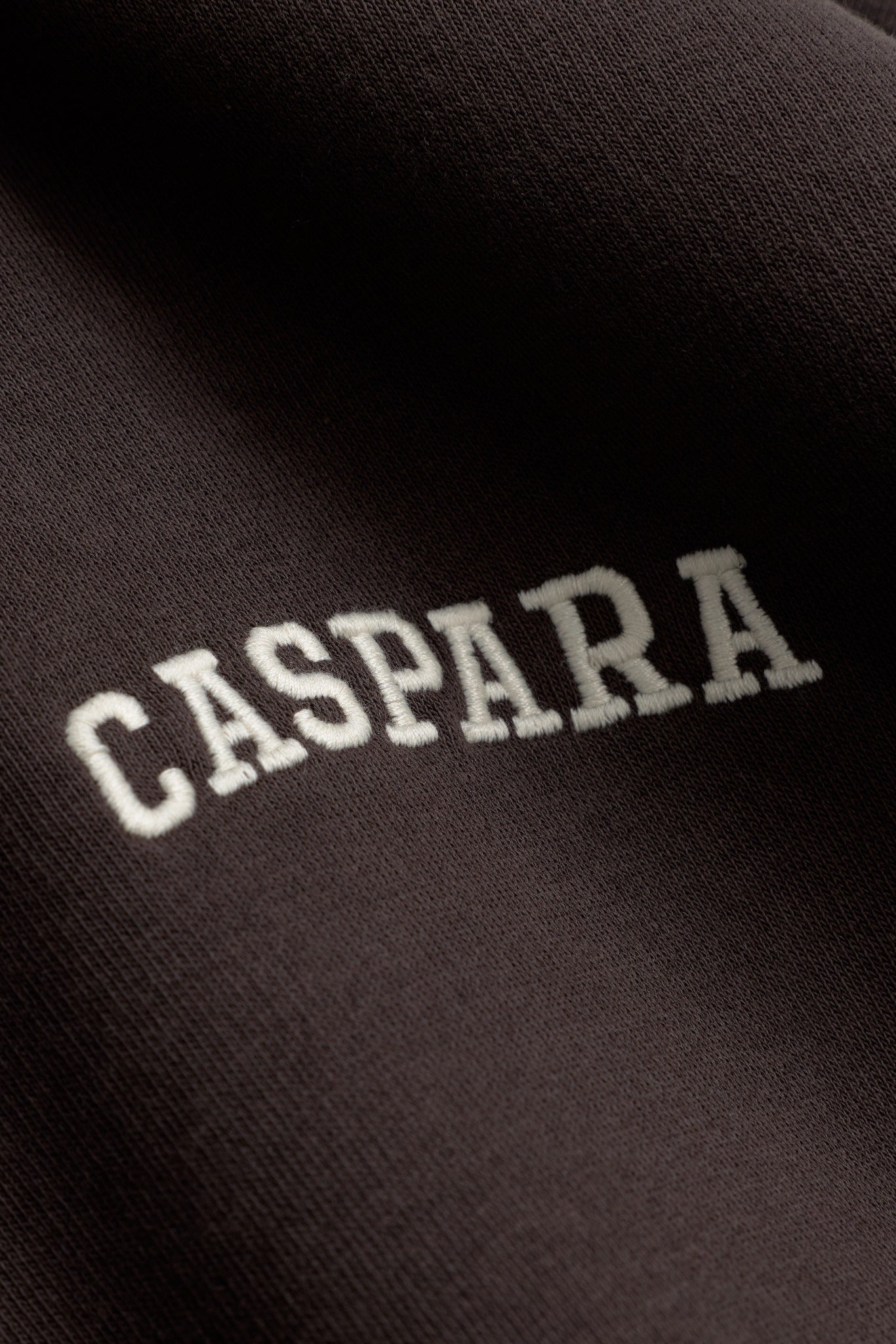 Close-up of the white embroidered CASPARA logo on espresso brown sweatshirt fabric.